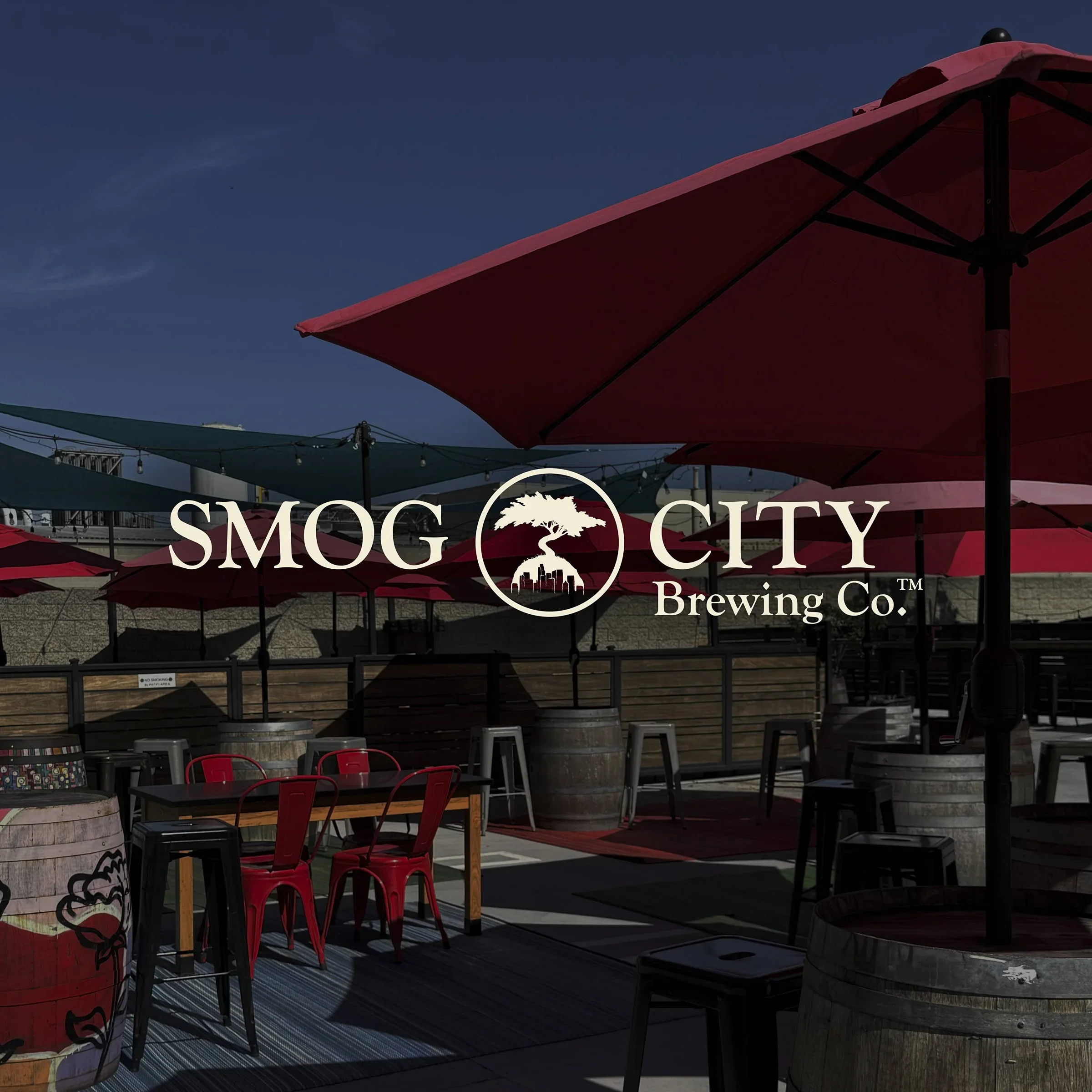 Outdoor seating area at Smog City Brewing Co. with red umbrellas, wooden tables, metal chairs, and barrels, under a clear blue sky.