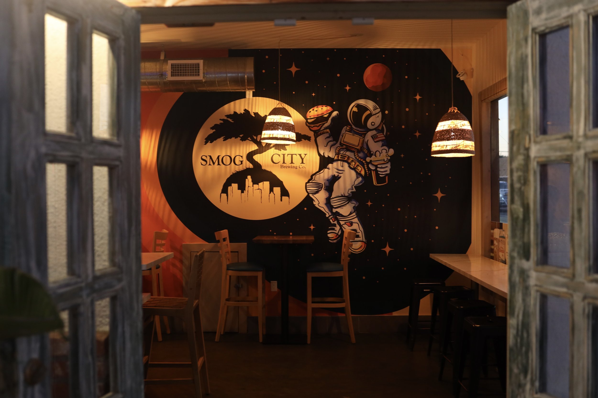 Interior of a brewery named Smog City Brewing Co. with space-themed wall art showing an astronaut holding a beer, a planet, stars, and a tree with city skyline on a black background, illuminated by hanging lamps.