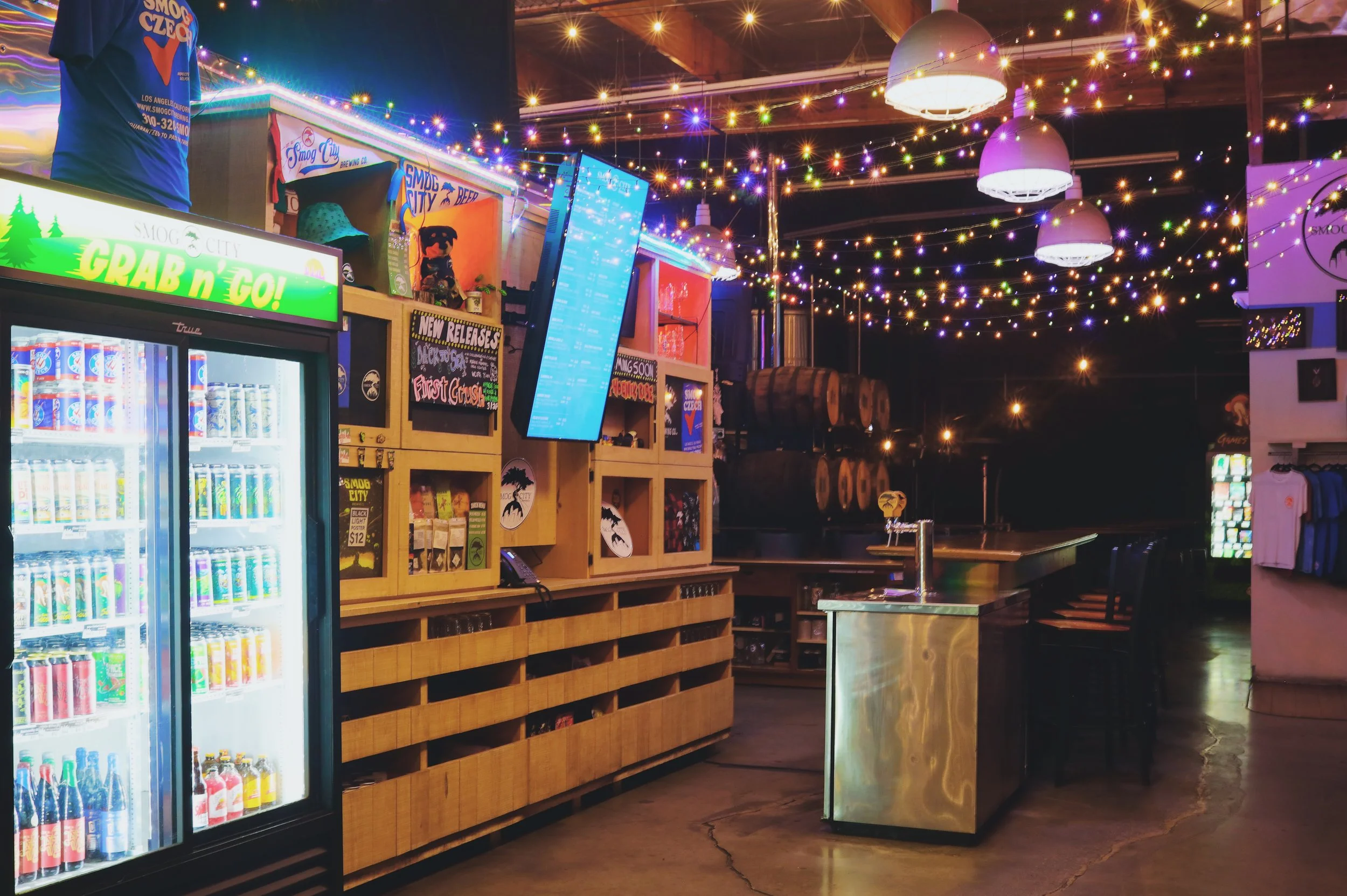 View of entrance to Smog City Brewery with counter, to-go fridge, digital beer menu, and overhead lights.