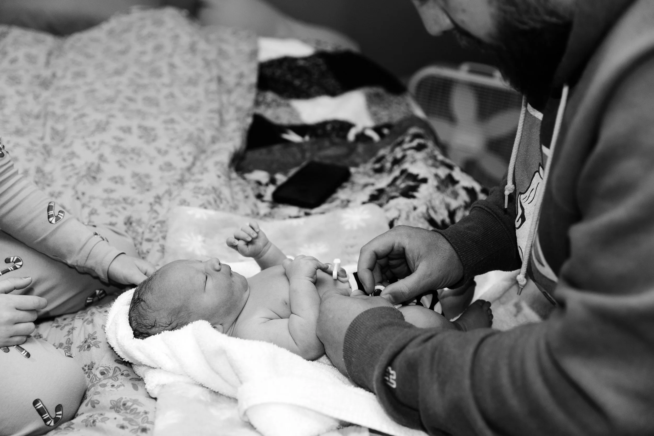 A newborn baby lying on a hospital bed holding a finger of an adult. The adult is standing next to the baby, and only their arm and hand are visible. The baby is wrapped in a towel or blanket, and the scene is in black and white.