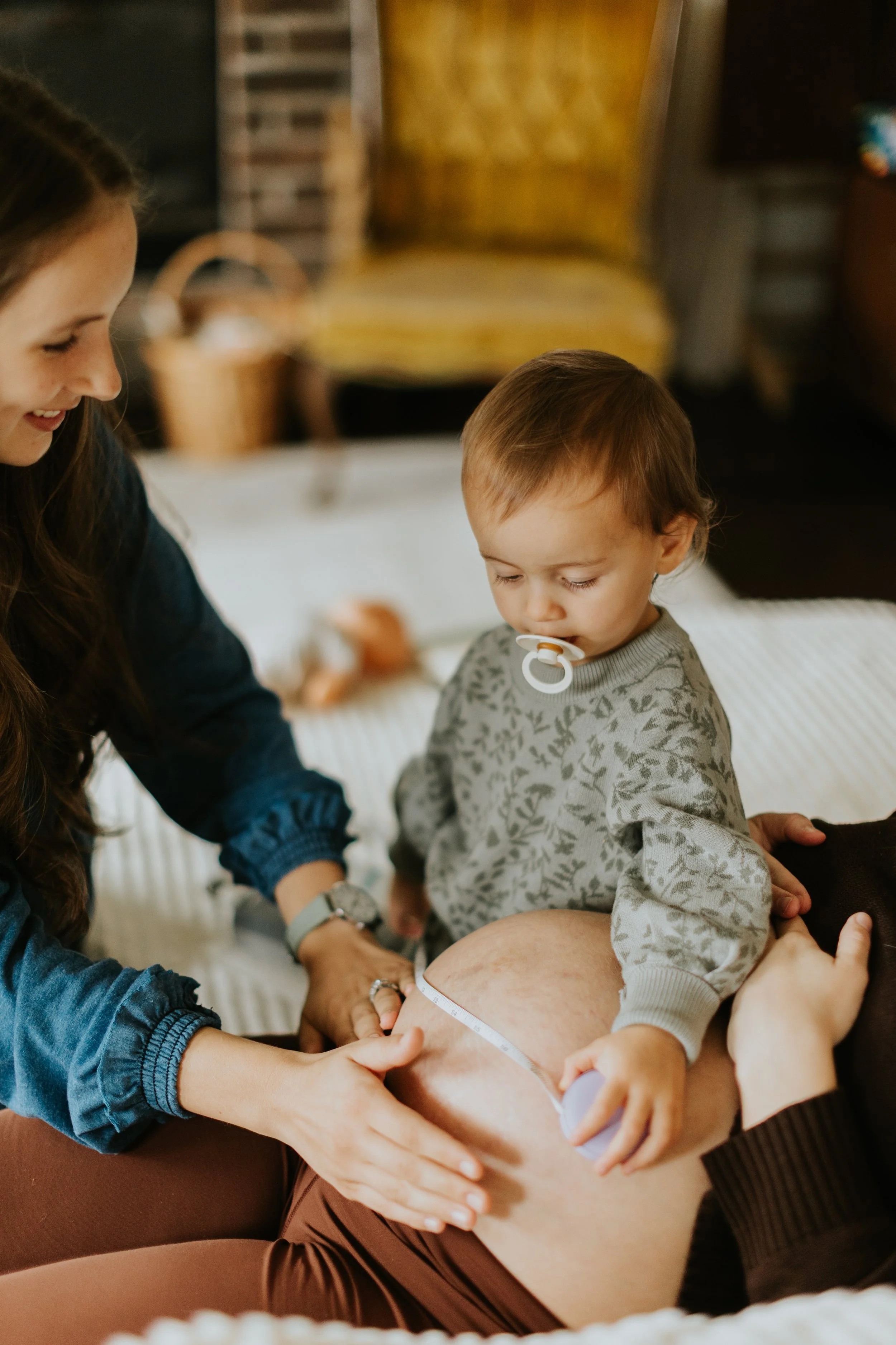 A midwife and older sibling measure a mother's belly while she lays on her cozy couch for a home prenatal visit.