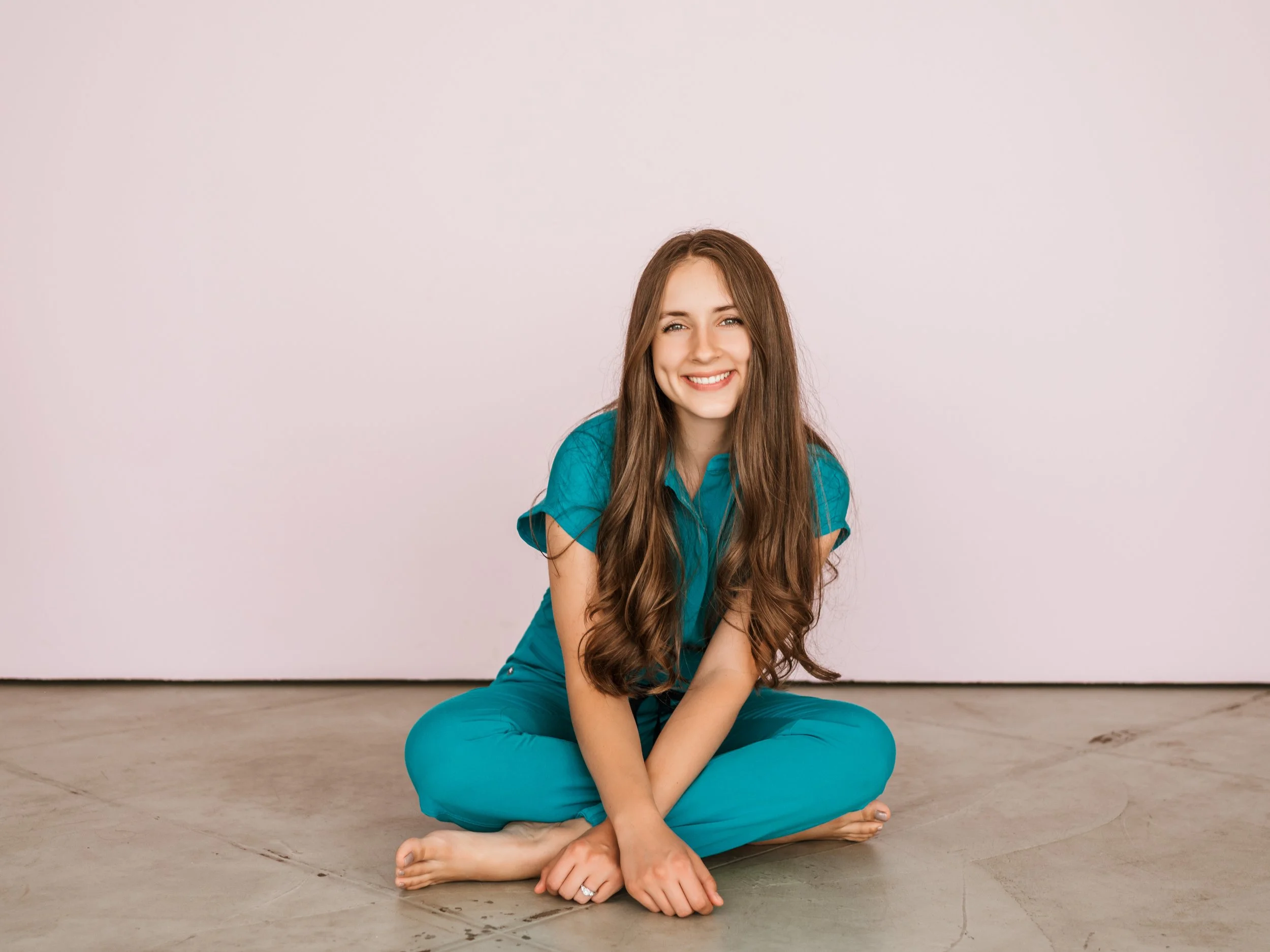 A young woman with long brown hair, wearing a teal shirt and pants, sitting on the floor cross-legged and smiling at the camera against a plain light-colored wall.