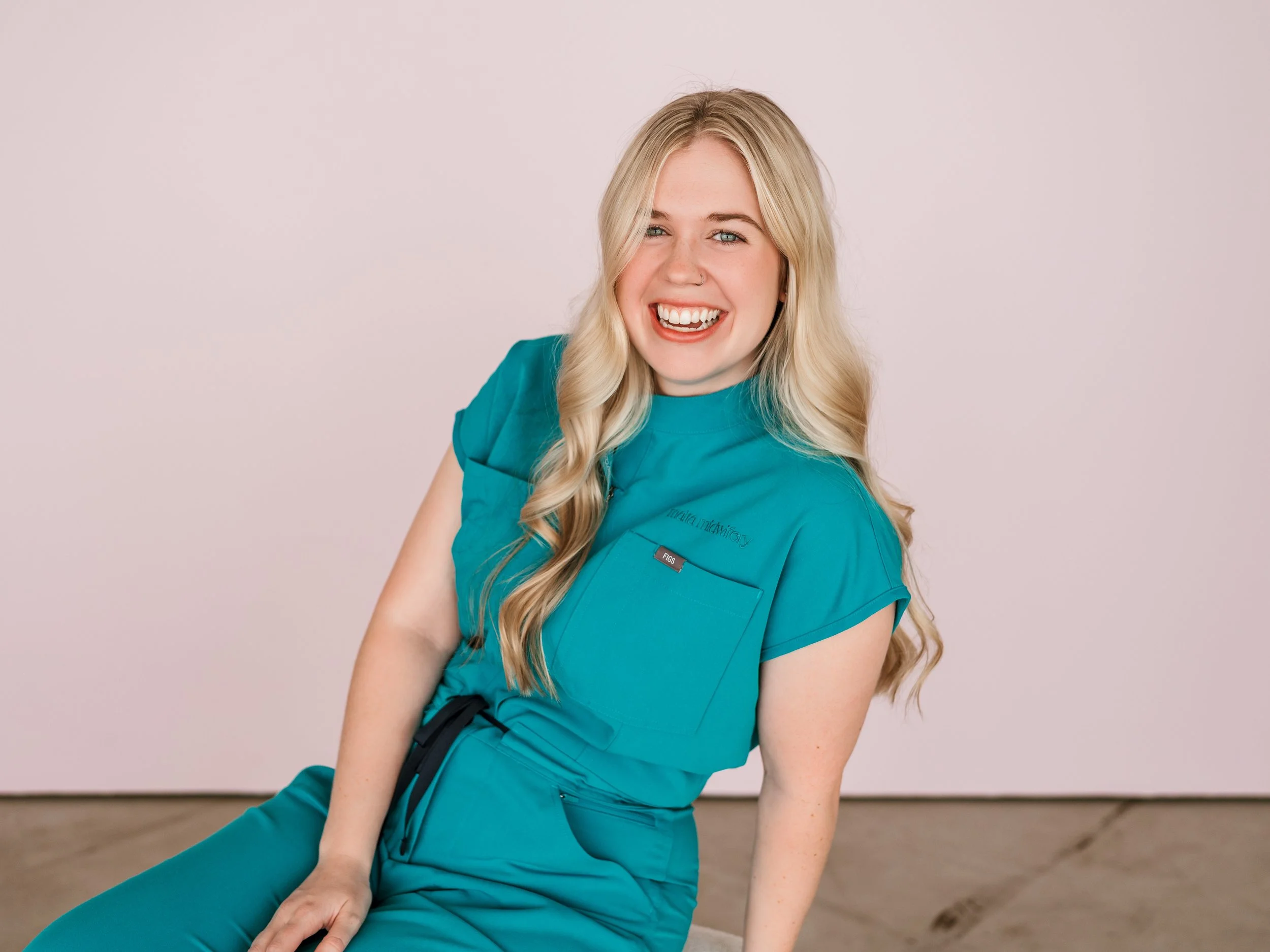 A young woman with long blonde hair wearing teal medical scrubs, smiling and sitting on the floor against a plain light pink background.