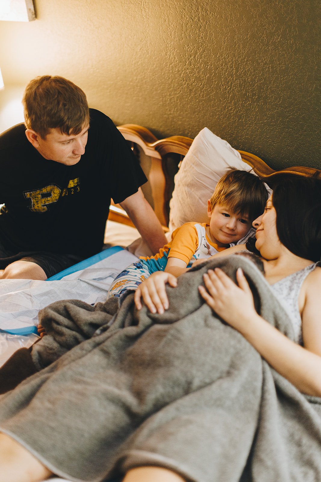 A family scene in a bedroom with a woman lying in bed, a young boy resting his head on her, and a man sitting on the bed, all engaging in a tender moment.