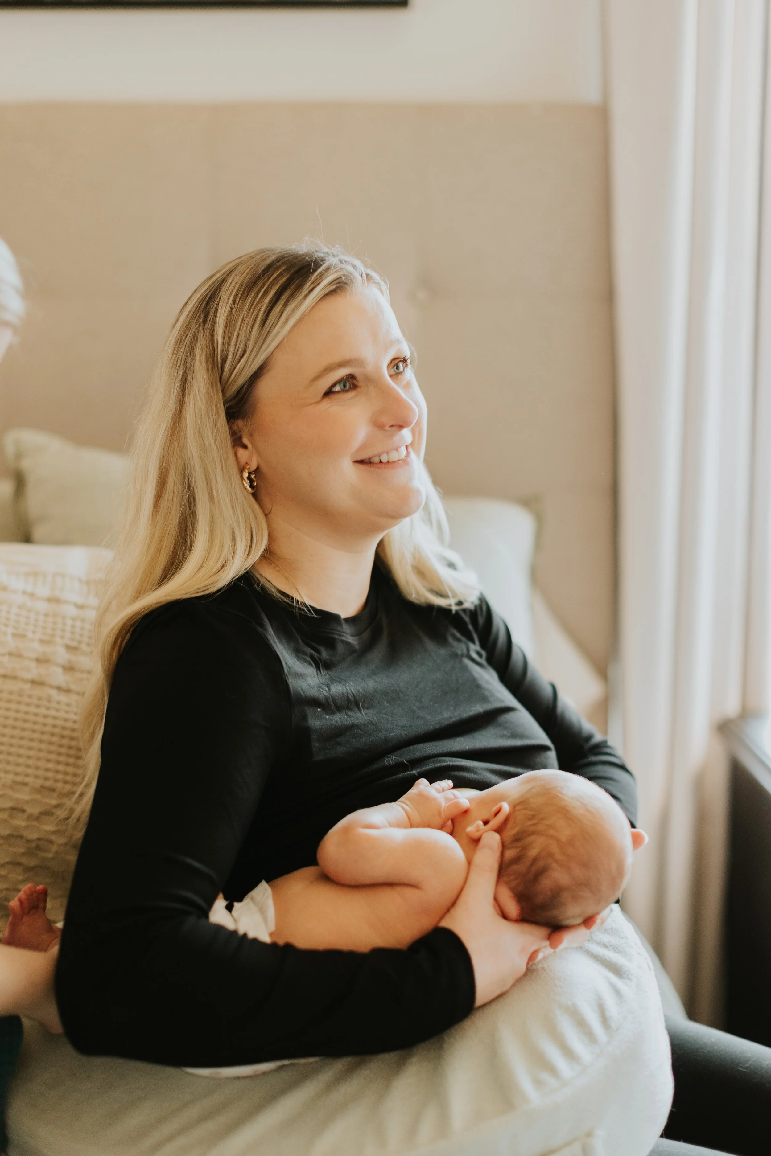 A woman with long blonde hair, wearing a black top, is sitting on a couch and smiling while holding a newborn baby against her chest near a window.