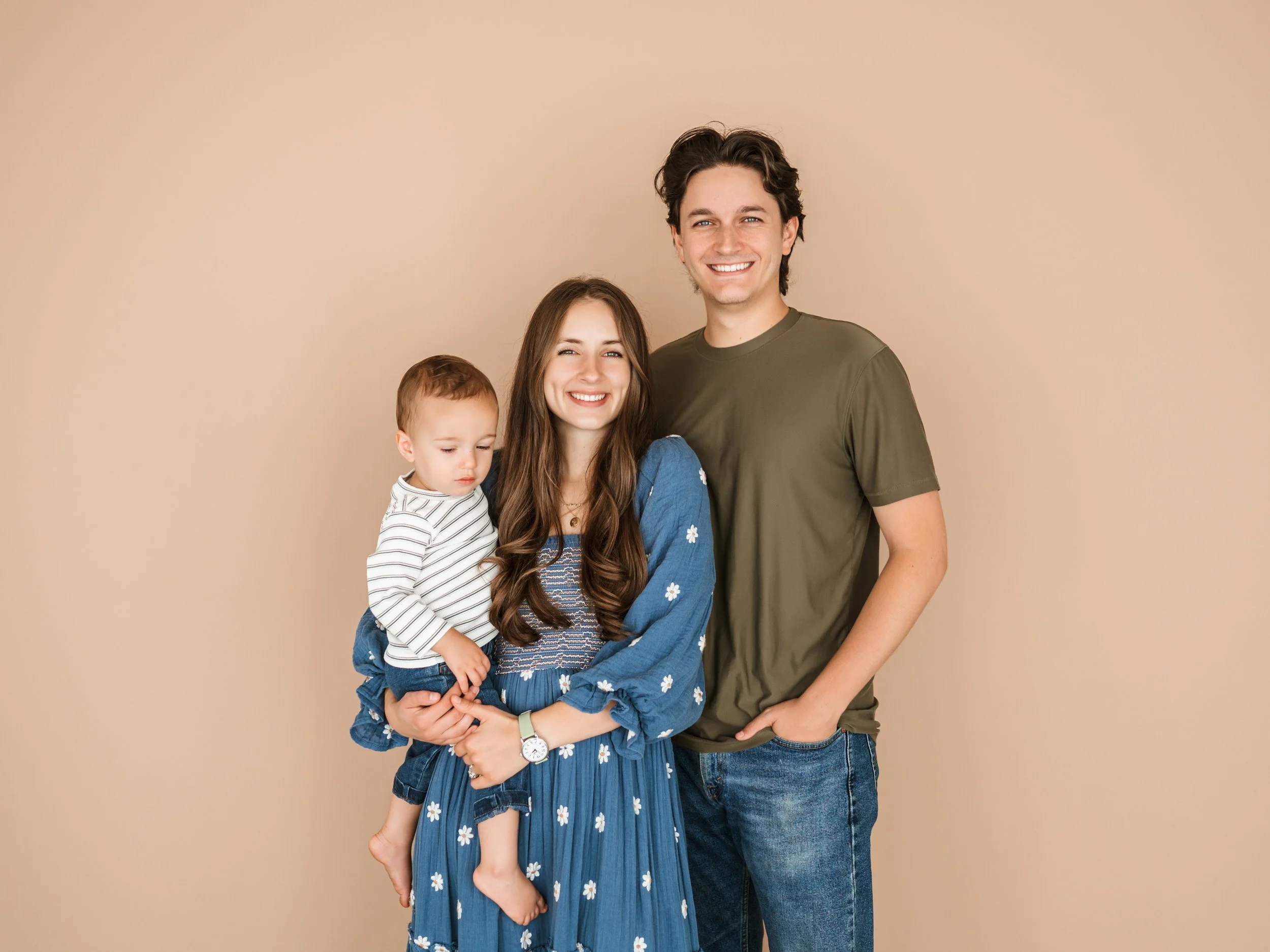 A smiling family of three posing against a beige background. A woman with long brown hair in a blue floral dress, a young child in a striped shirt held by the woman, and a man with dark hair in a green t-shirt and jeans.