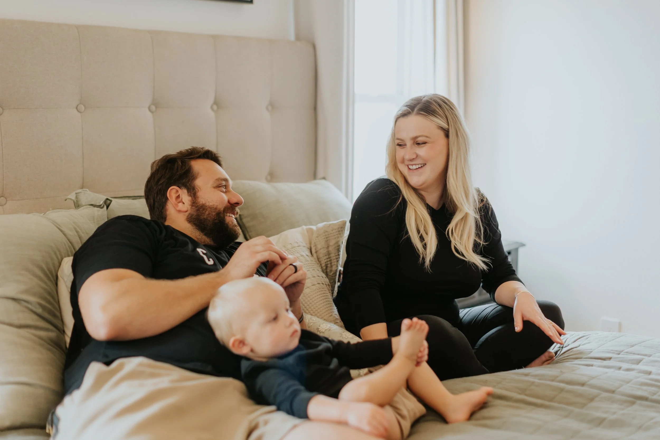 A family of three sitting on a bed, smiling and engaging with each other. The man is lying down, the woman is sitting, and a young child is sitting on the bed.