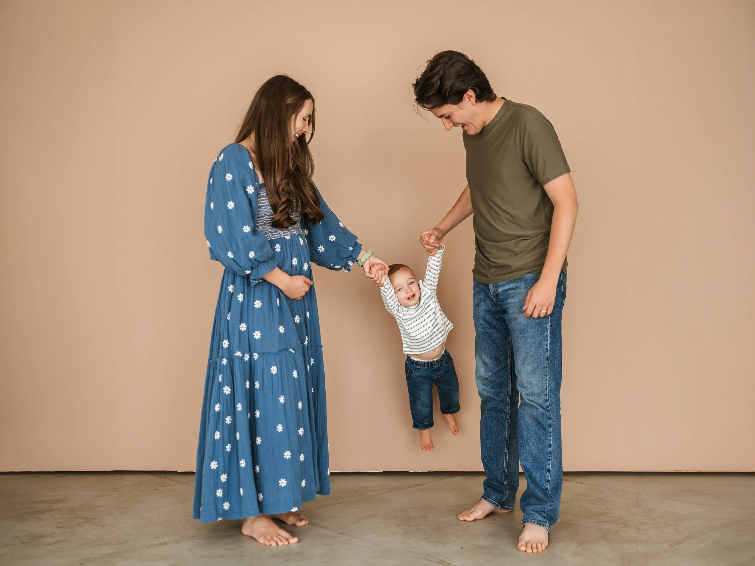 A young family of three holding hands and playing together indoors against a beige background. The mother and father are each holding one of their smiling child's hands, who is hanging mid-air in the middle.