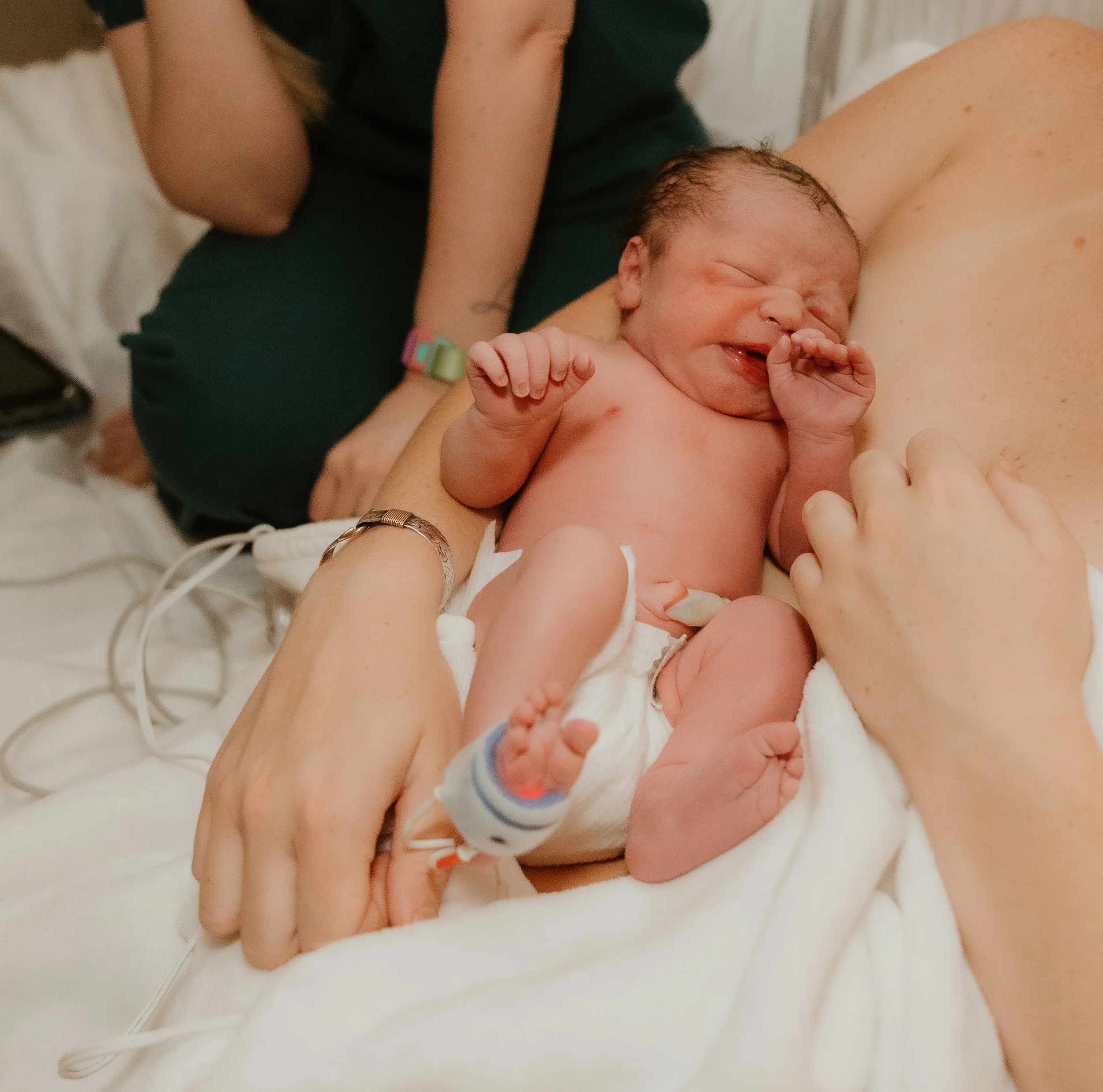 A newborn baby with closed eyes, smiling, lying on an adult's arm, with a pulse oximeter on her foot, and part of the adult's hand gently holding the baby's arm.