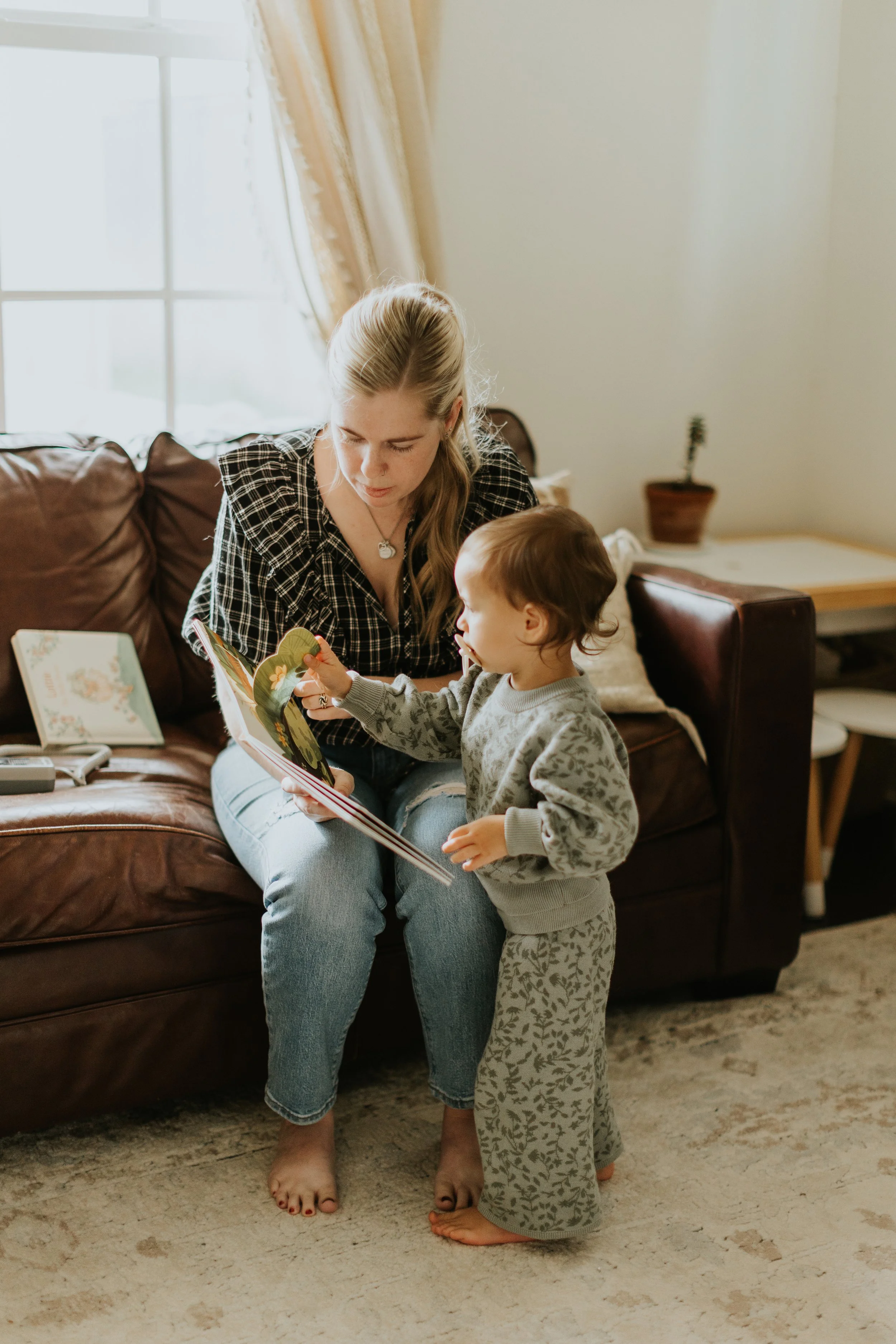 A home birth midwife sits on a couch in a client's home a reads a book to their toddler.