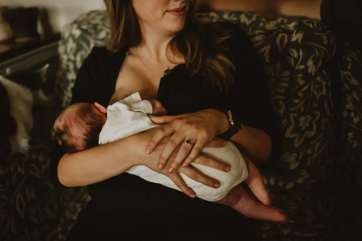 A woman in a black top holding a newborn baby on her chest, sitting on a patterned sofa in a dimly lit room.