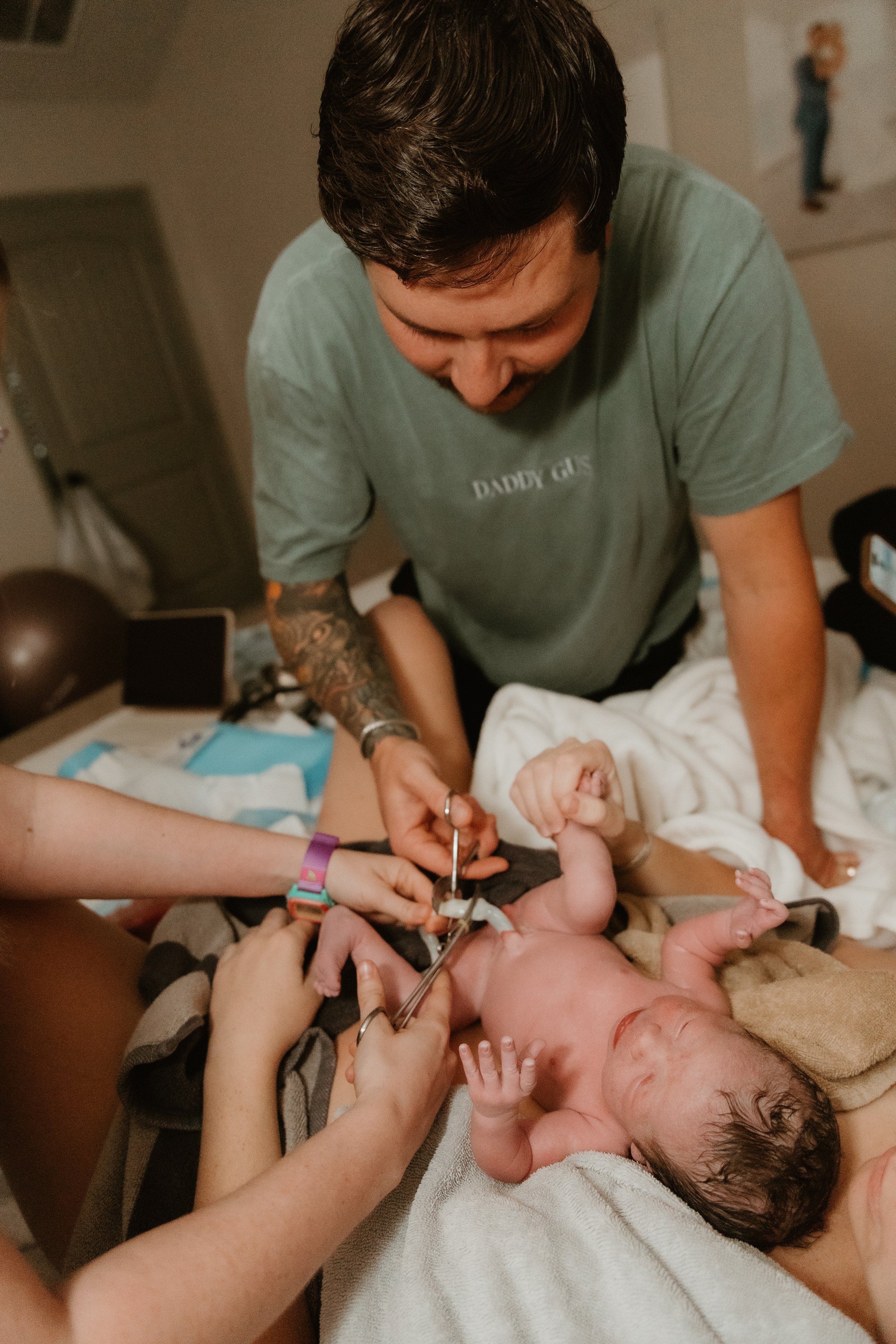 A woman and a man are delivering a baby with a midwife at home. A father cuts the umbilical cord. 