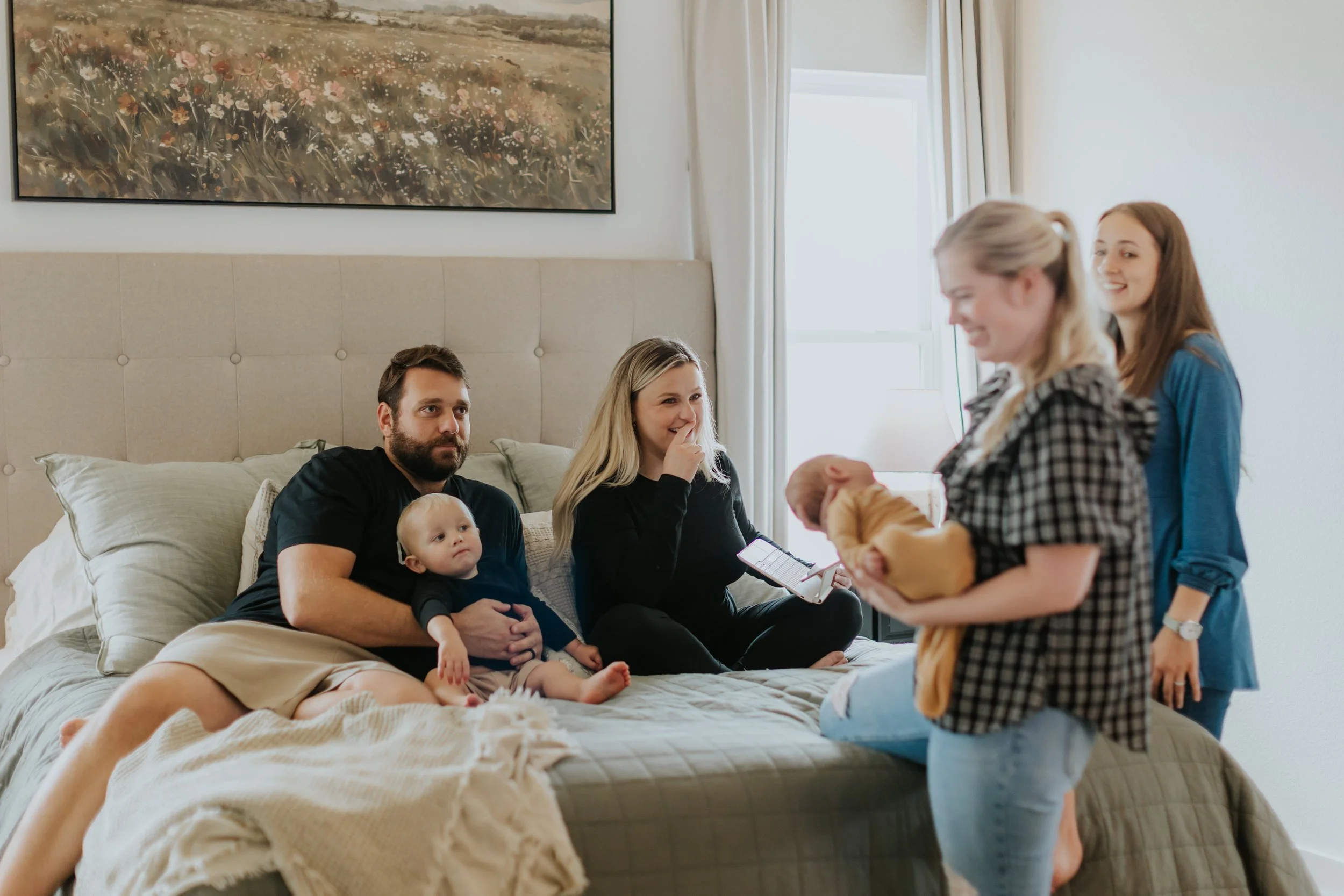 Family gathering in a bedroom with five women, a man, and two babies. One woman is holding a baby, and two women are standing near the bed. The man and woman are sitting on the bed, smiling and engaging with the person standing.