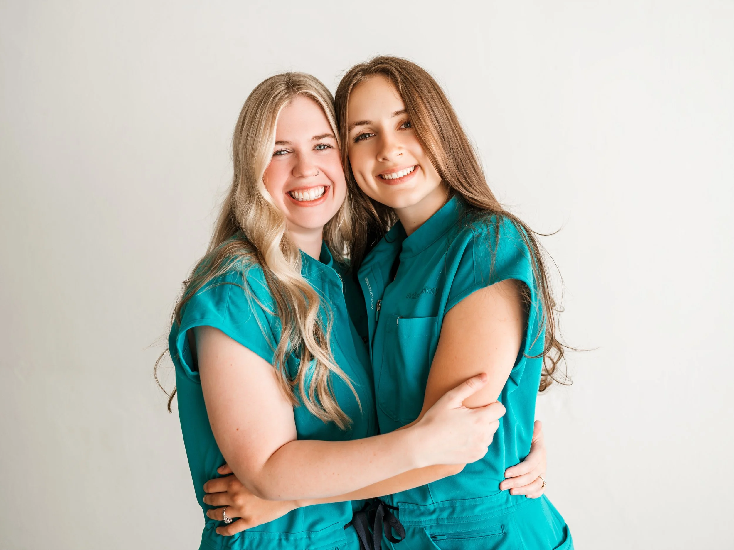 Two women in teal scrubs smiling and hugging each other against a white background.