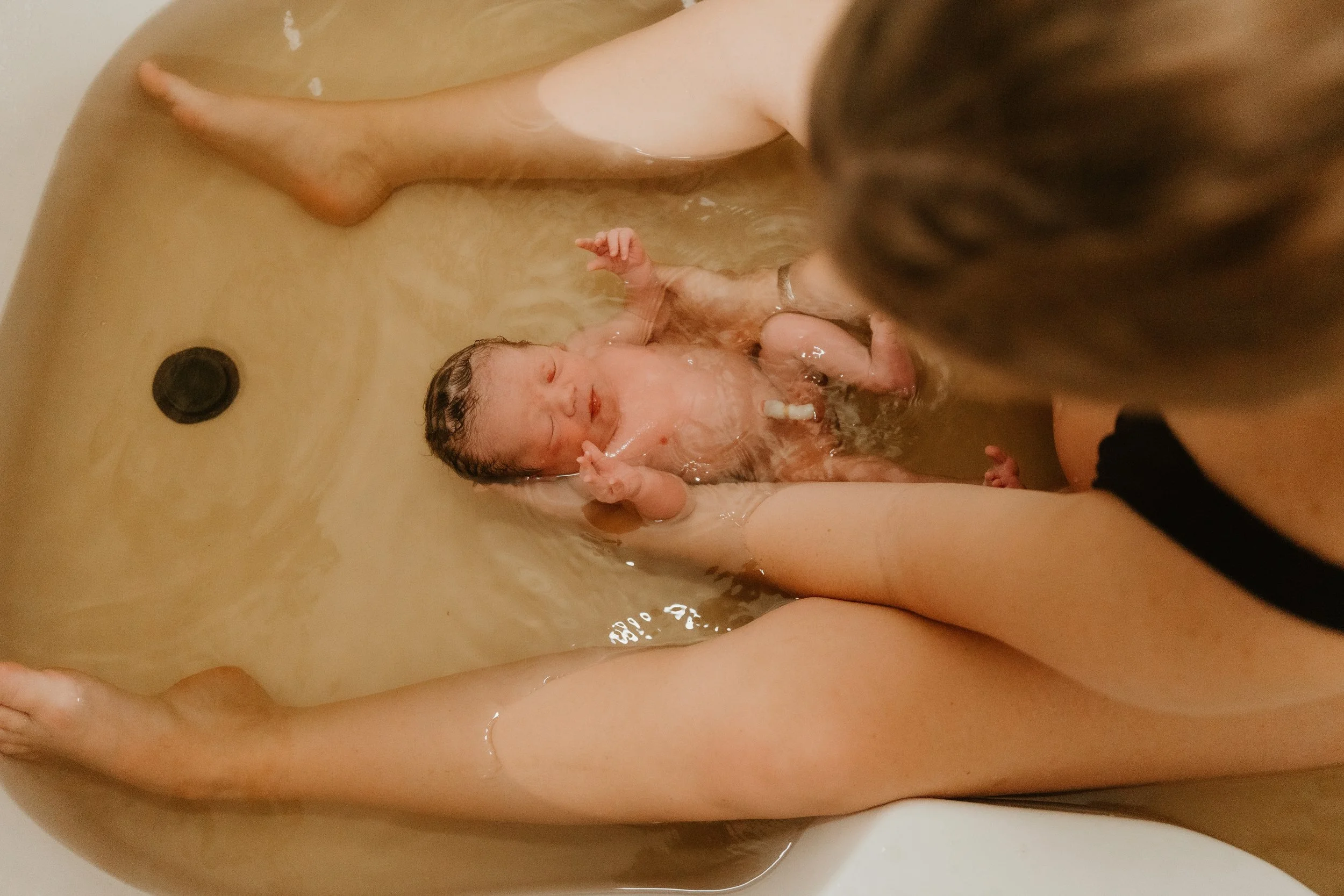 A newborn baby in a bathtub with water, being supported by an adult's hands during a bath.