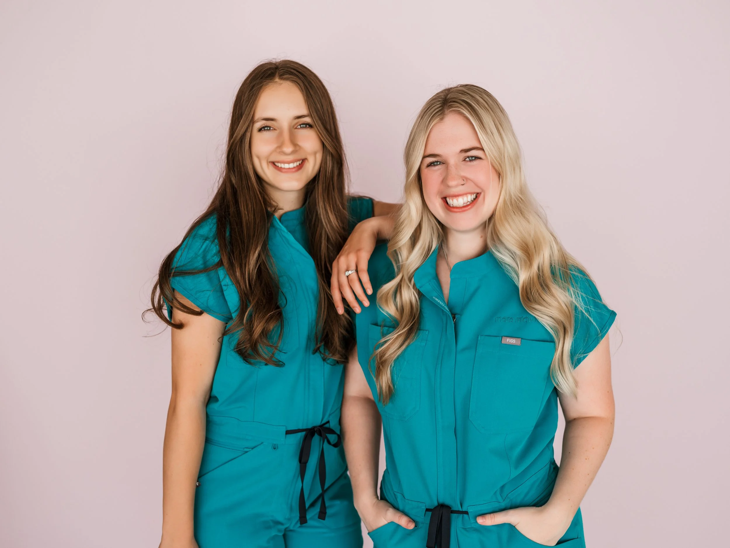 Two women in turquoise medical scrubs smiling, one with brown hair and one with blonde hair, standing against a light background.