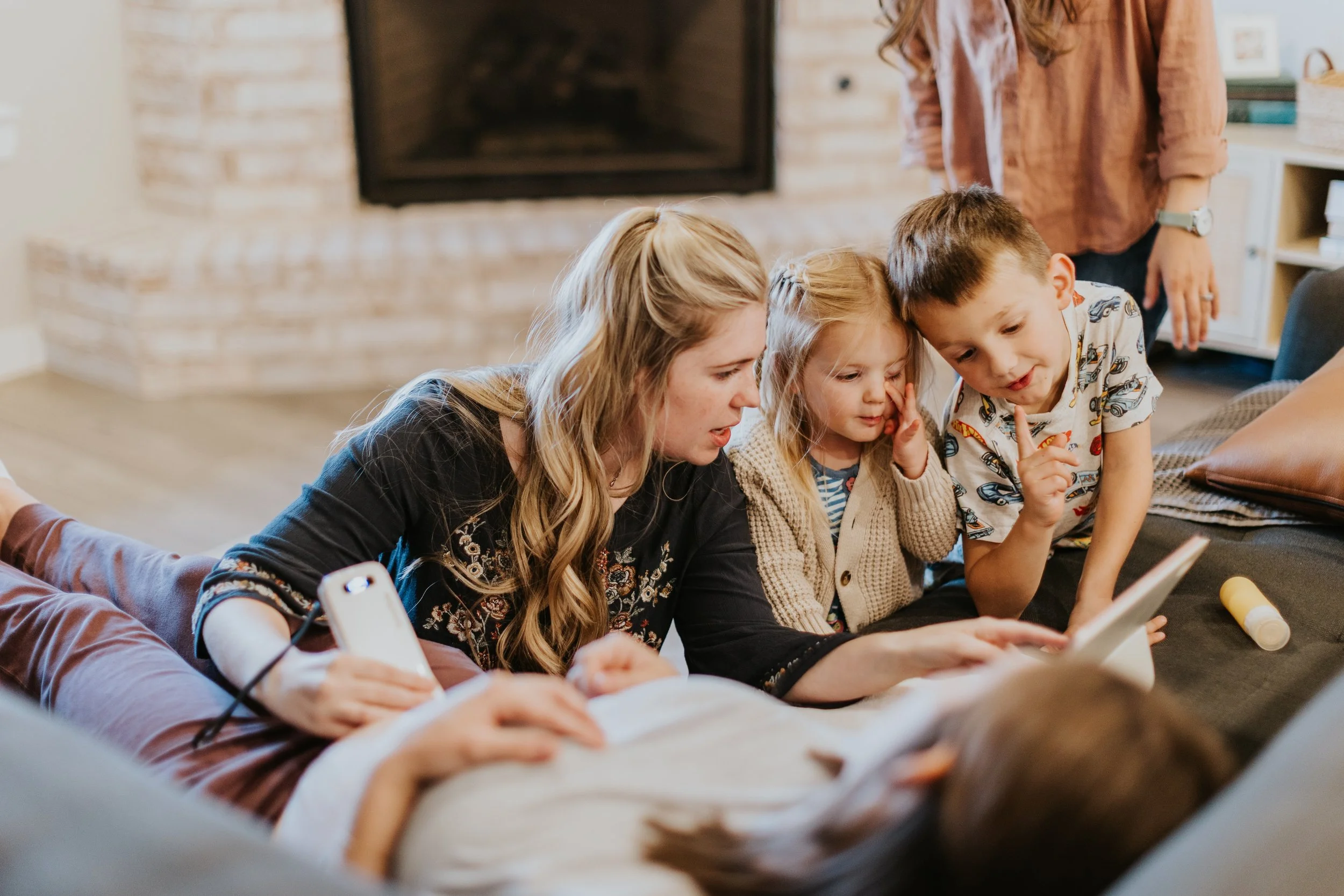 A group of four people, including two adults and two children, are lying on the floor together, looking at a tablet device. They are in a cozy living room with a brick fireplace in the background.