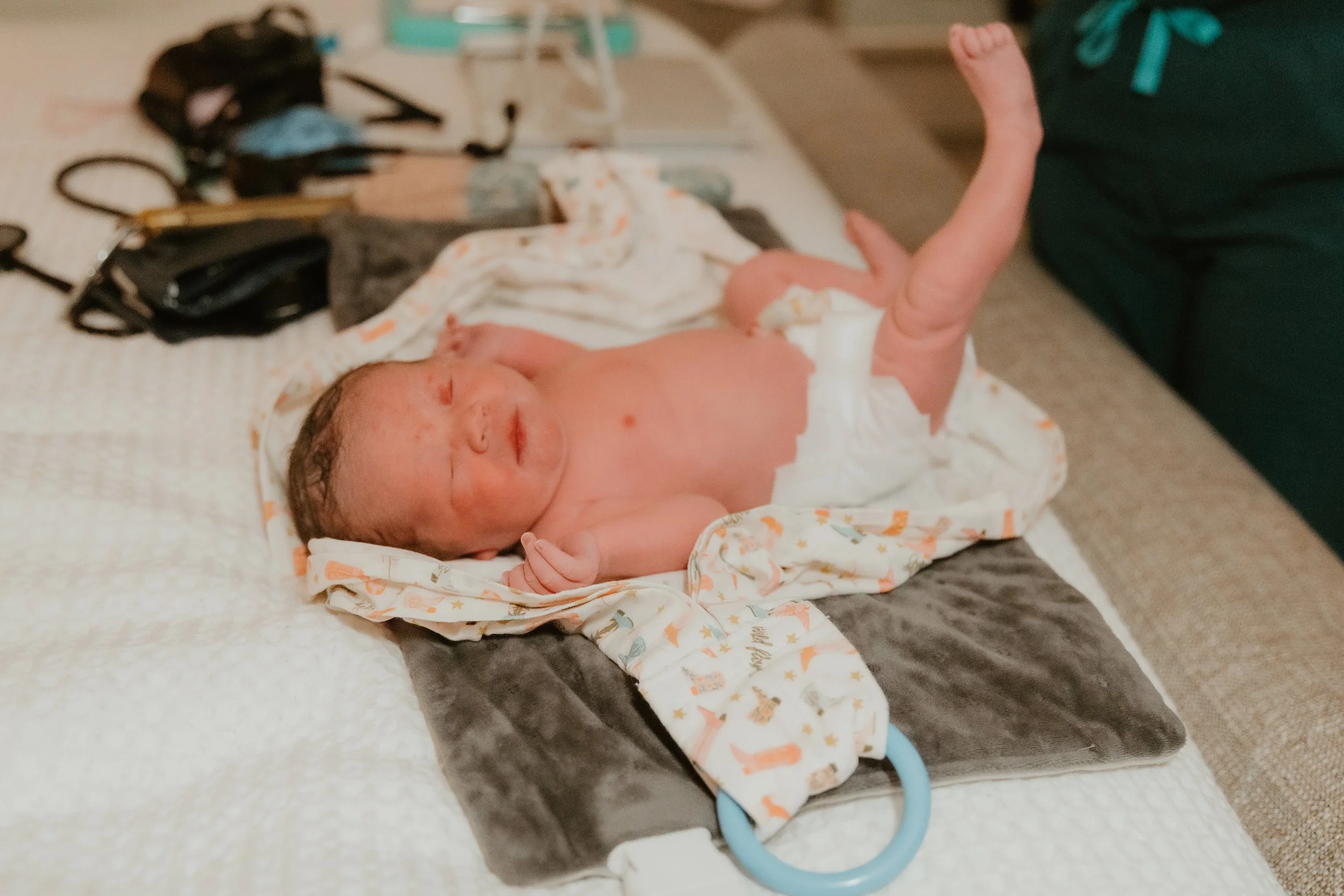 Newborn baby lying on hospital bassinet, wrapped in a blanket, with medical equipment and supplies in the background.