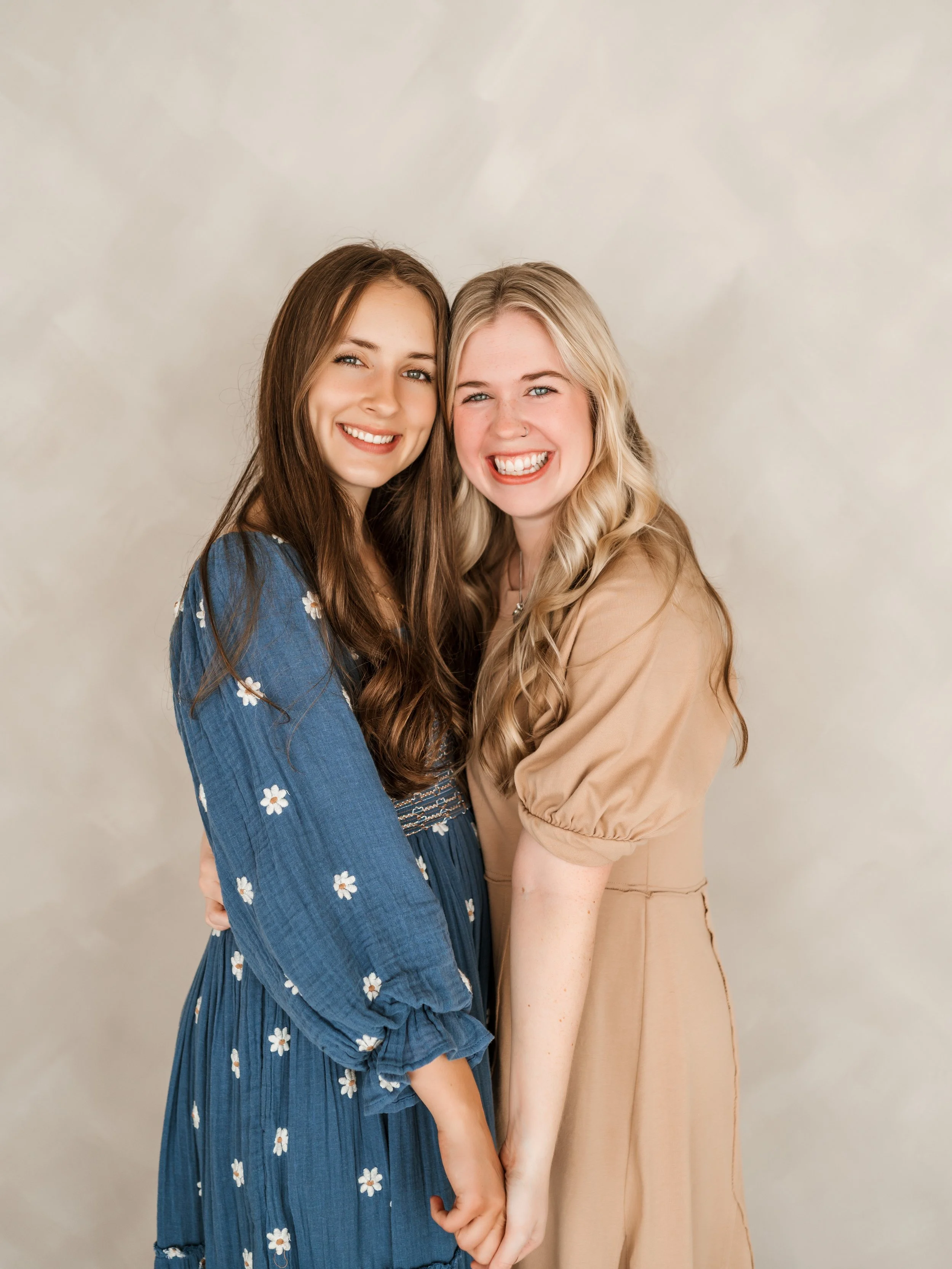 Two young women smiling and holding hands, standing close together against a neutral background.