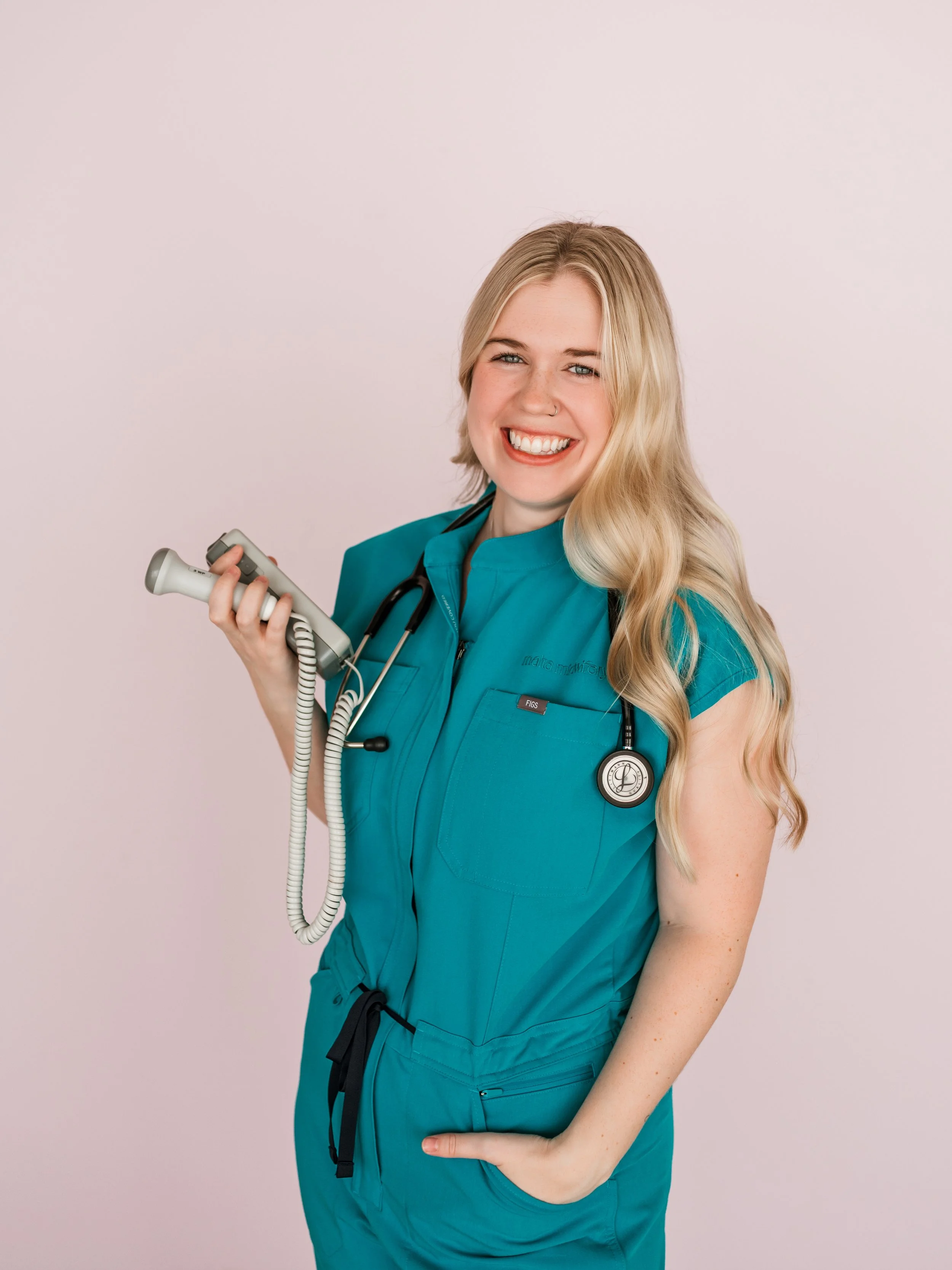 A smiling female healthcare professional with long blonde hair, wearing teal scrubs with a stethoscope around her neck, holding a medical device, standing against a plain background.