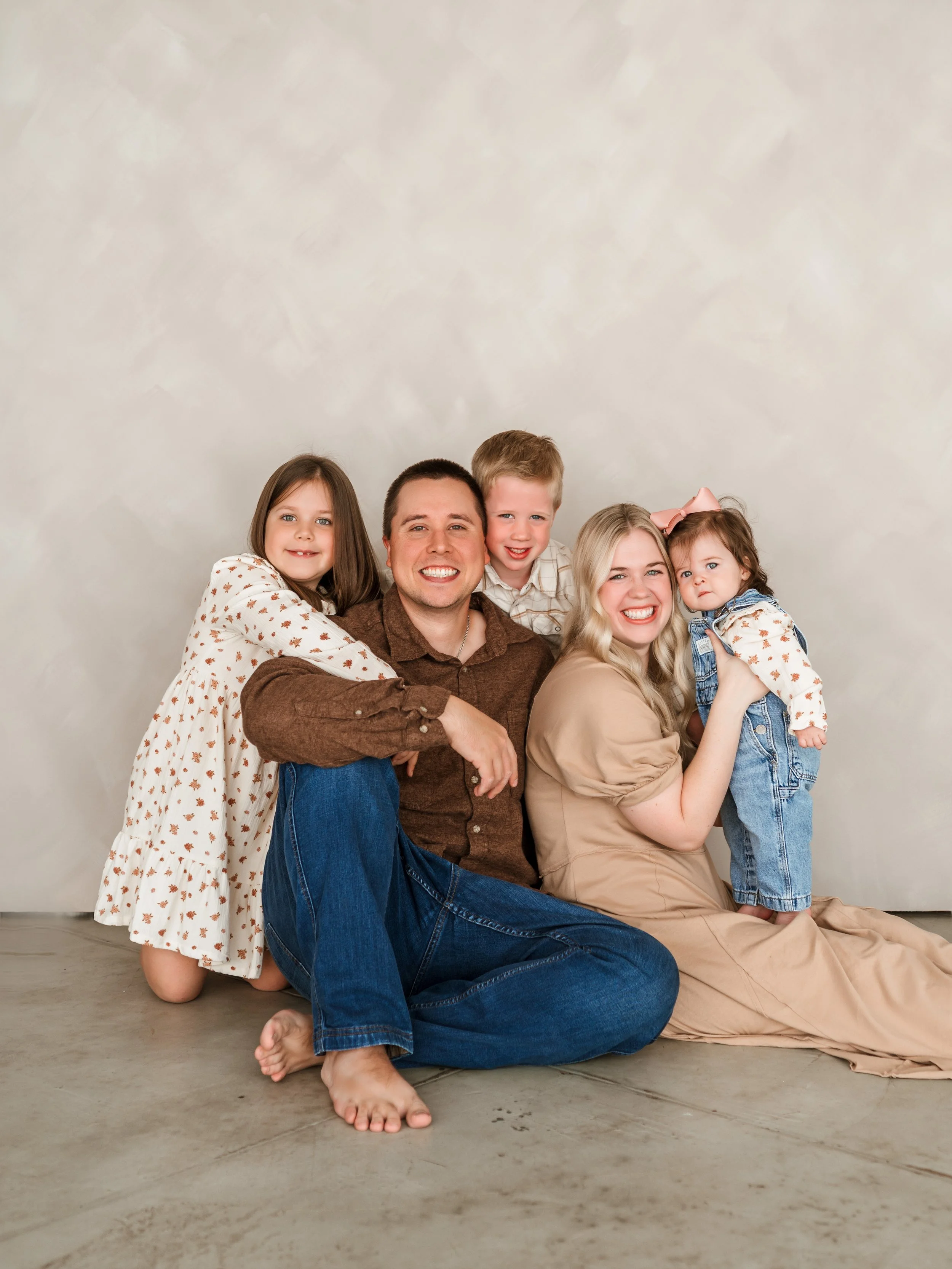A smiling family of five sitting on the floor, with a beige background. The family includes a man, woman, and three children, all smiling and interacting with each other.