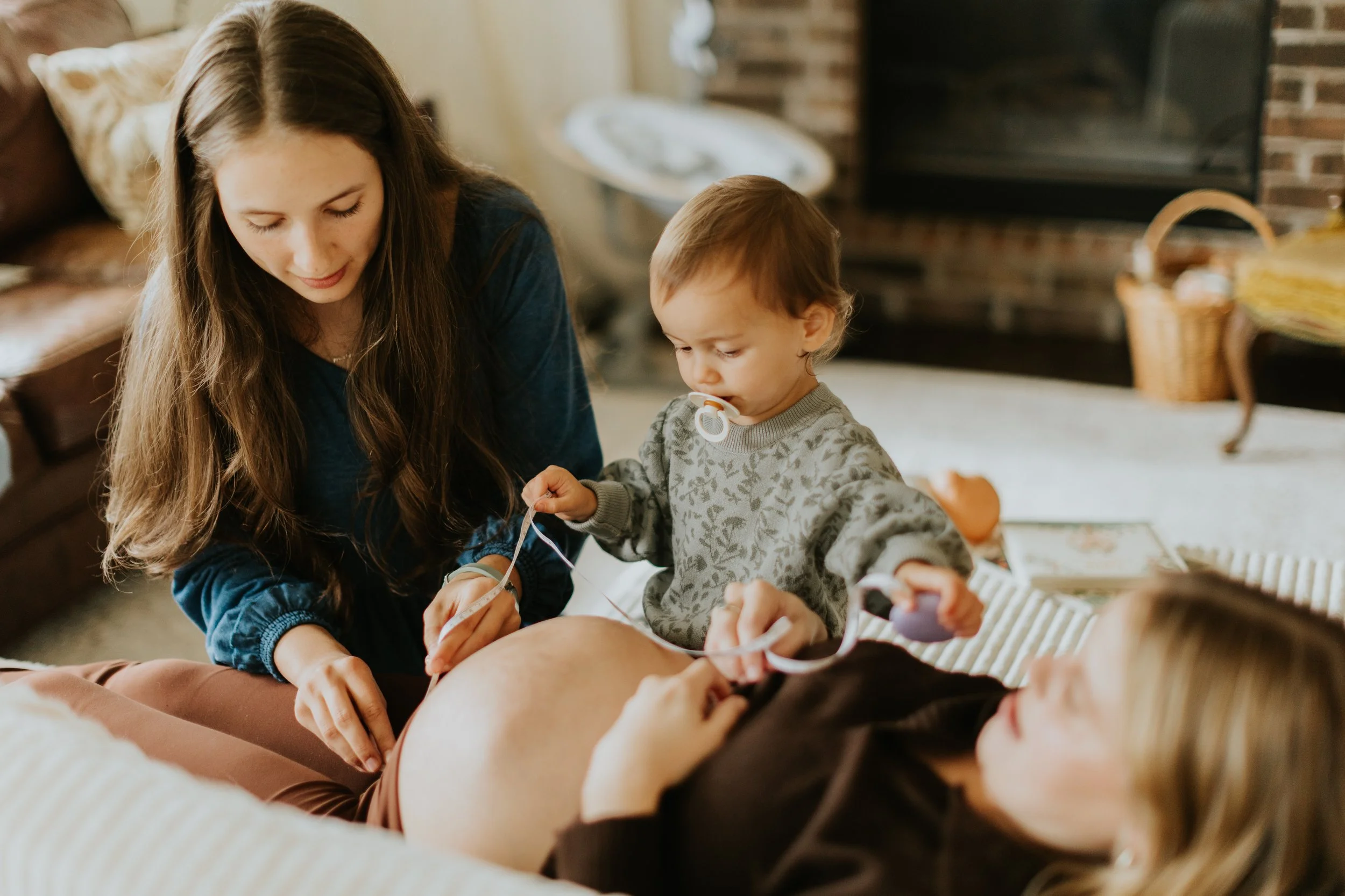 A woman lying on her back on a couch with a pregnant belly while a midwife and older child measure her pregnant belly in a cozy living room.