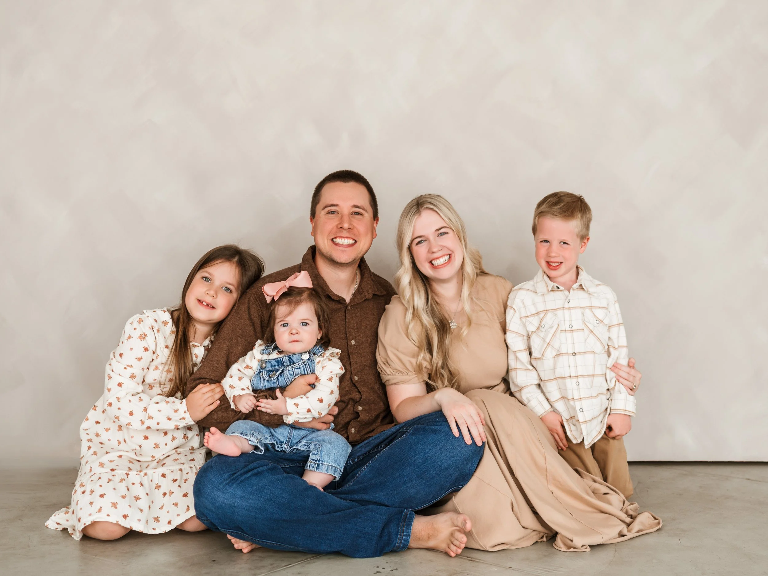 Family portrait of six people, two adults and four children, sitting and smiling on the floor against a neutral background, dressed in casual and coordinated clothing.