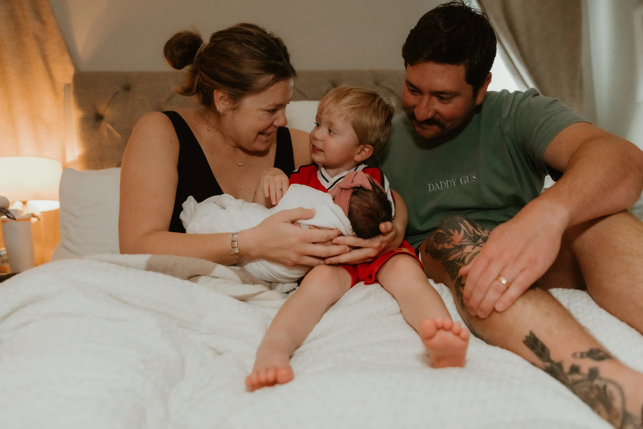 A family of four, including a woman, man, young boy, and newborn baby, sitting on a bed and smiling at each other in a warmly lit bedroom.