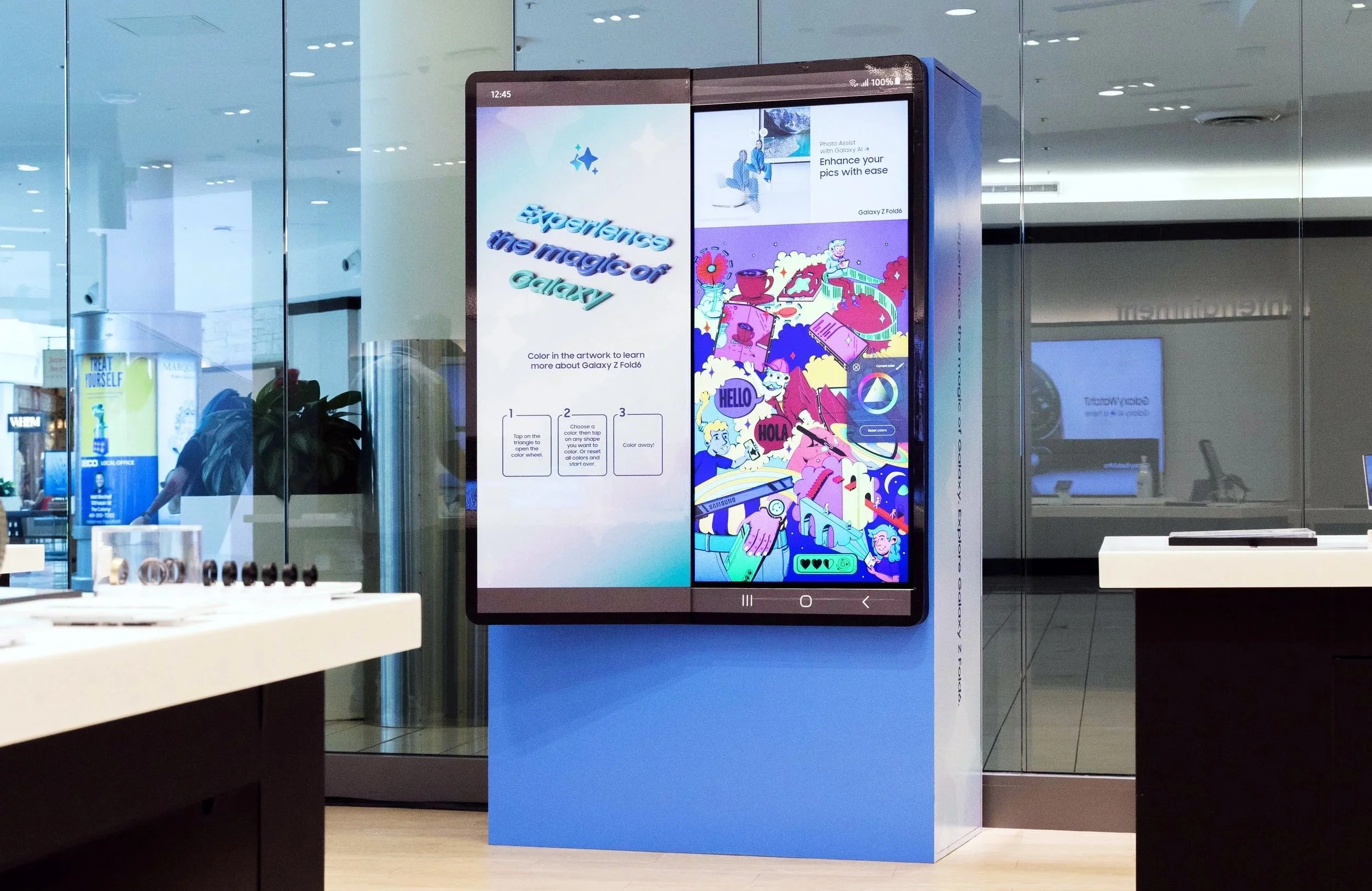 Digital display screen in a shopping mall or store, showing colorful artwork and instructions for coloring to learn about the Galaxy Z Fold6. The display is mounted on a blue stand, with reflections of the mall environment and other digital screens visible.