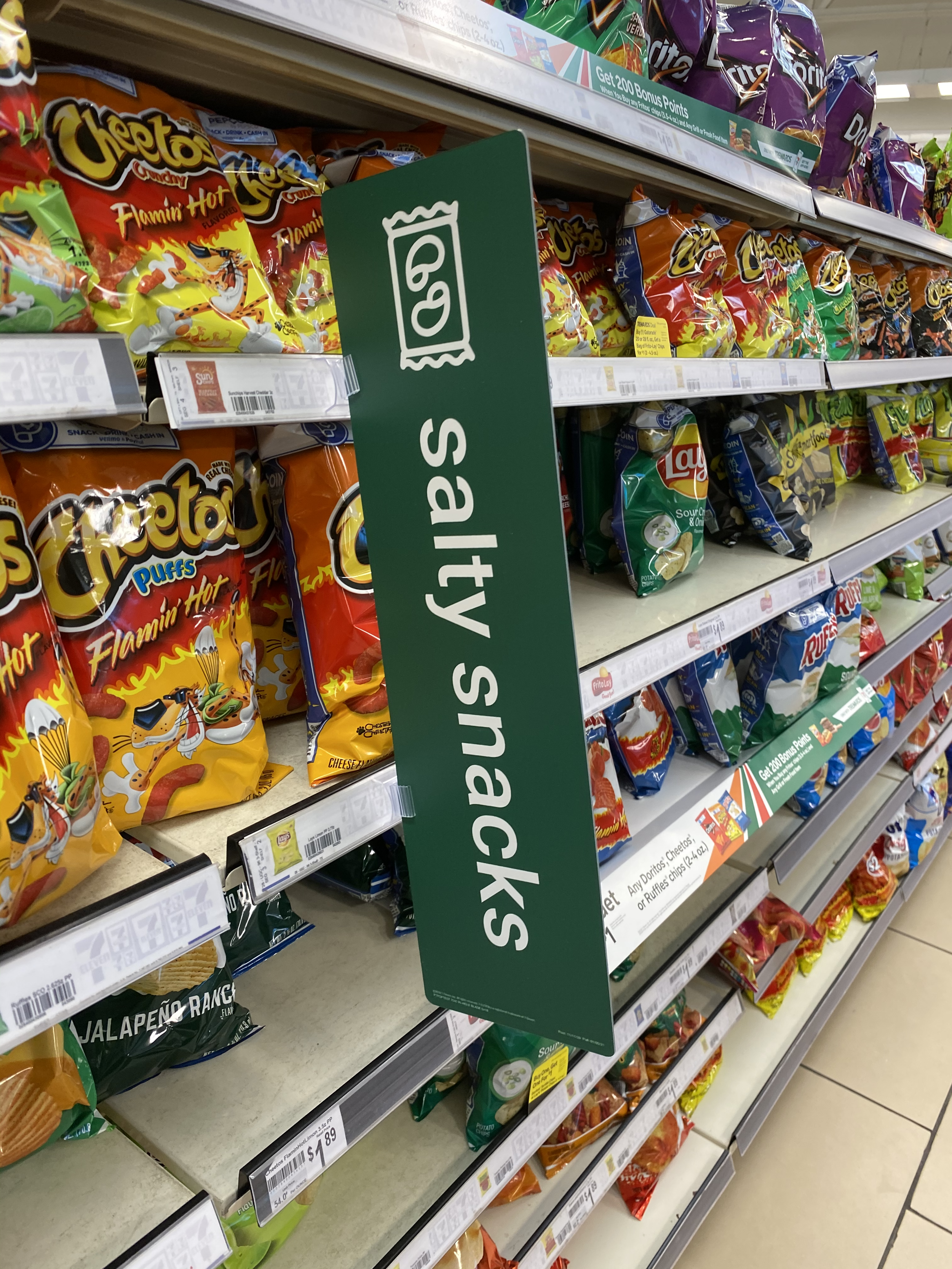 Supermarket aisle with bags of chips, including Cheetos Flamin' Hot and Lay's sour cream and onion, with a green 'Salty Snacks' sign hanging from the shelf.