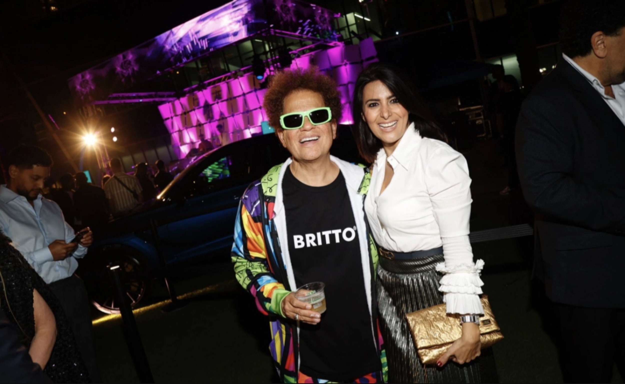 Two women posing at an outdoor event during nighttime, one with multicolored jacket and green sunglasses, holding a drink, and the other in a white blouse with a golden clutch, both smiling. Bright purple and pink lights illuminate the background.