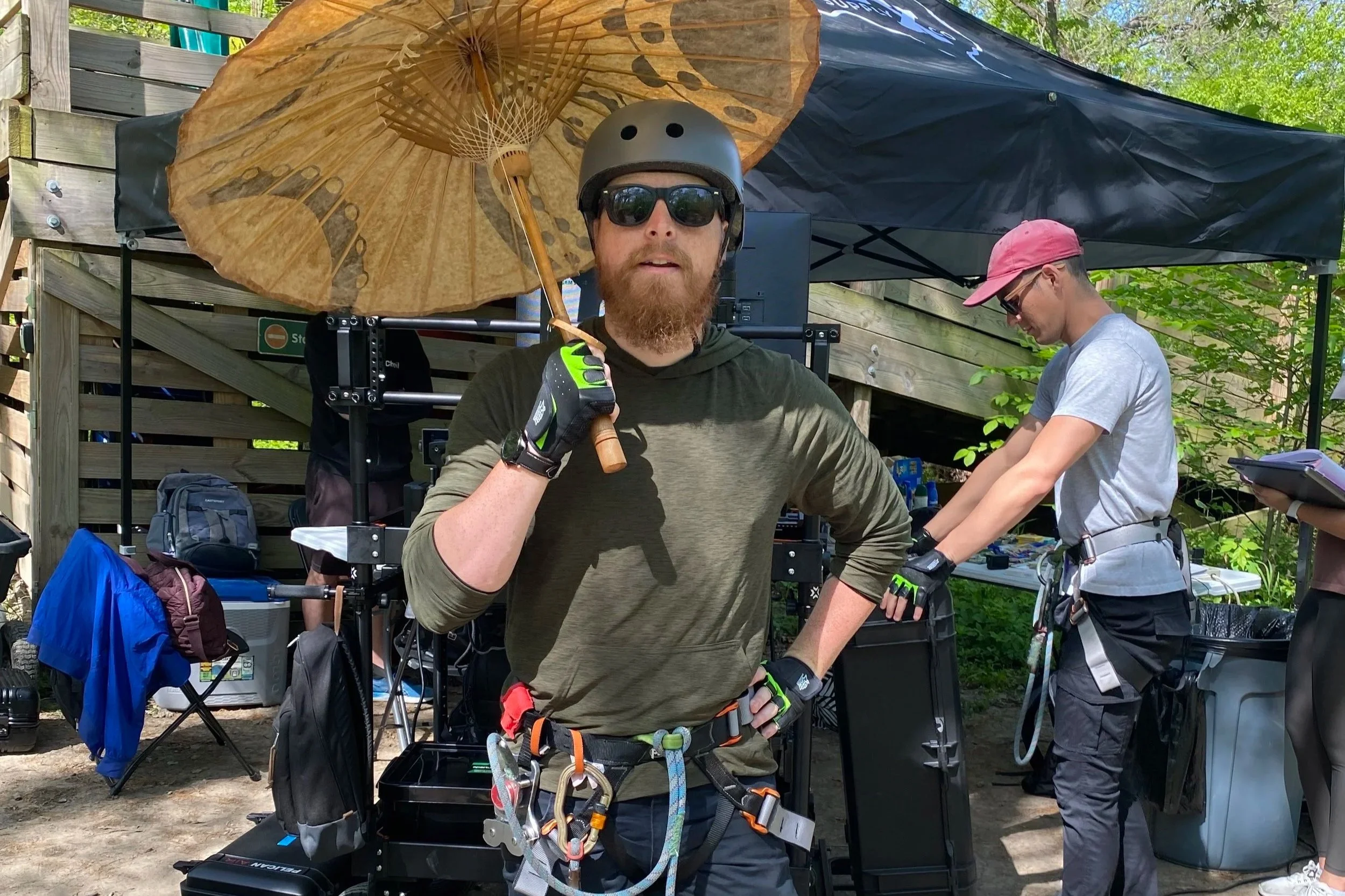 A man wearing a helmet, sunglasses, and climbing harness stands under a parasol in an outdoor setting, with equipment and another person working nearby.