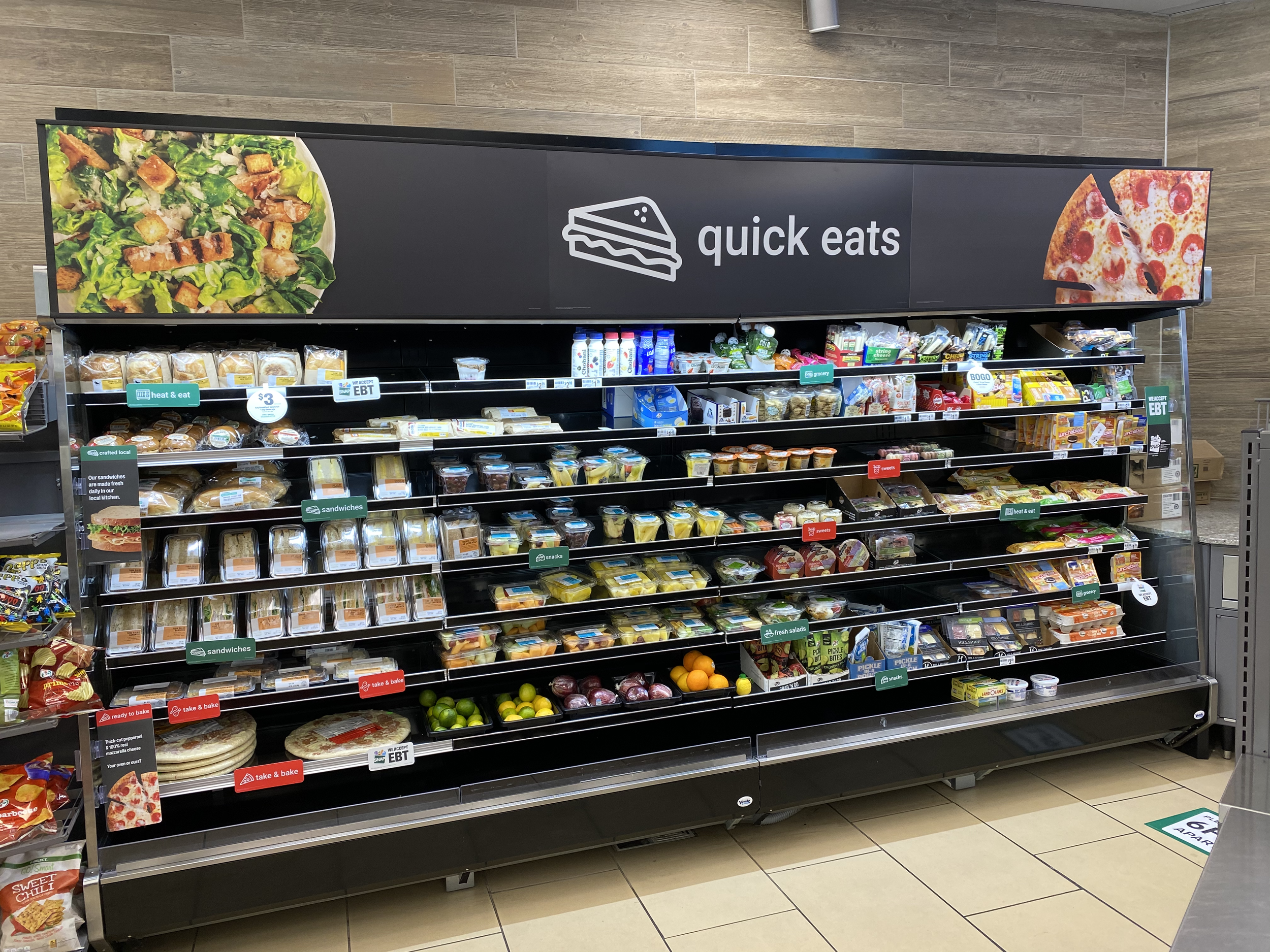Grocery store self-service refrigerator shelf stocked with pre-packaged salads, sandwiches, yogurt, fruits, and other ready-to-eat foods. Signage indicates categories such as salads, sandwiches, snacks, and baked items. The display is labeled 'quick eats' with images of a salad and pizza.