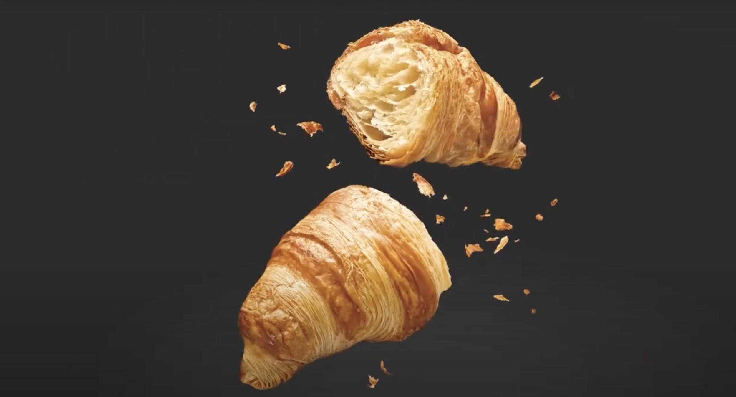 A croissant breaking in half with crumbs flying, on a black background.