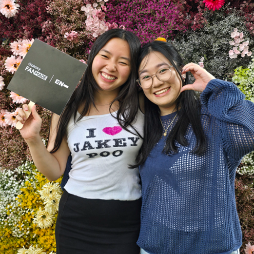 Two young women smiling and posing together in front of a colorful flower wall, one holding a Galaxy FanMade box.