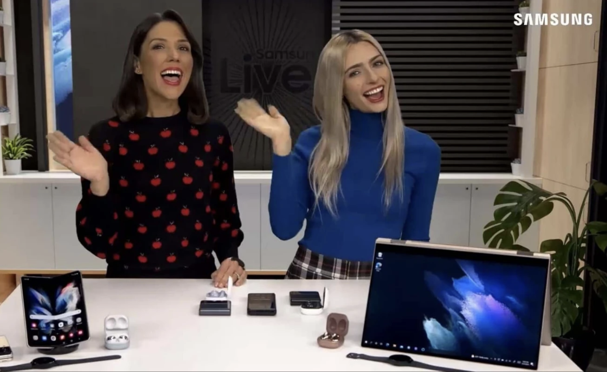 Two women smiling and waving in an office with Samsung devices and gadgets on the table in front of them.