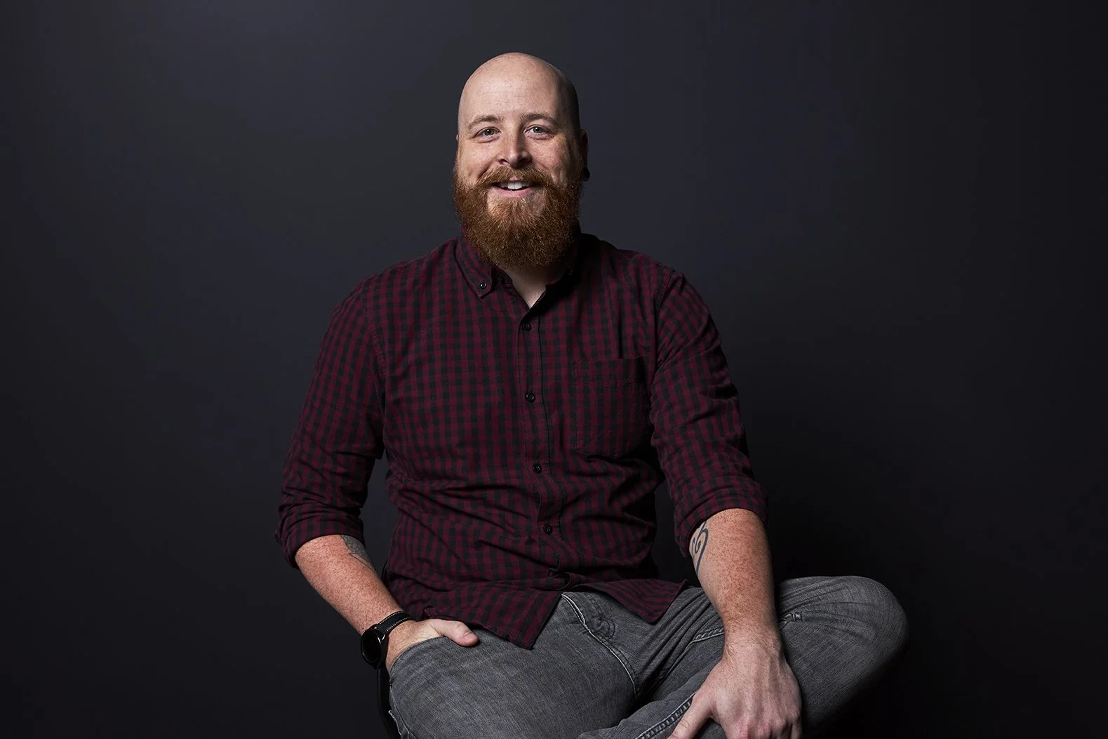 A man with a beard and mustache, wearing a red and black checkered shirt and gray jeans, sitting against a dark background.