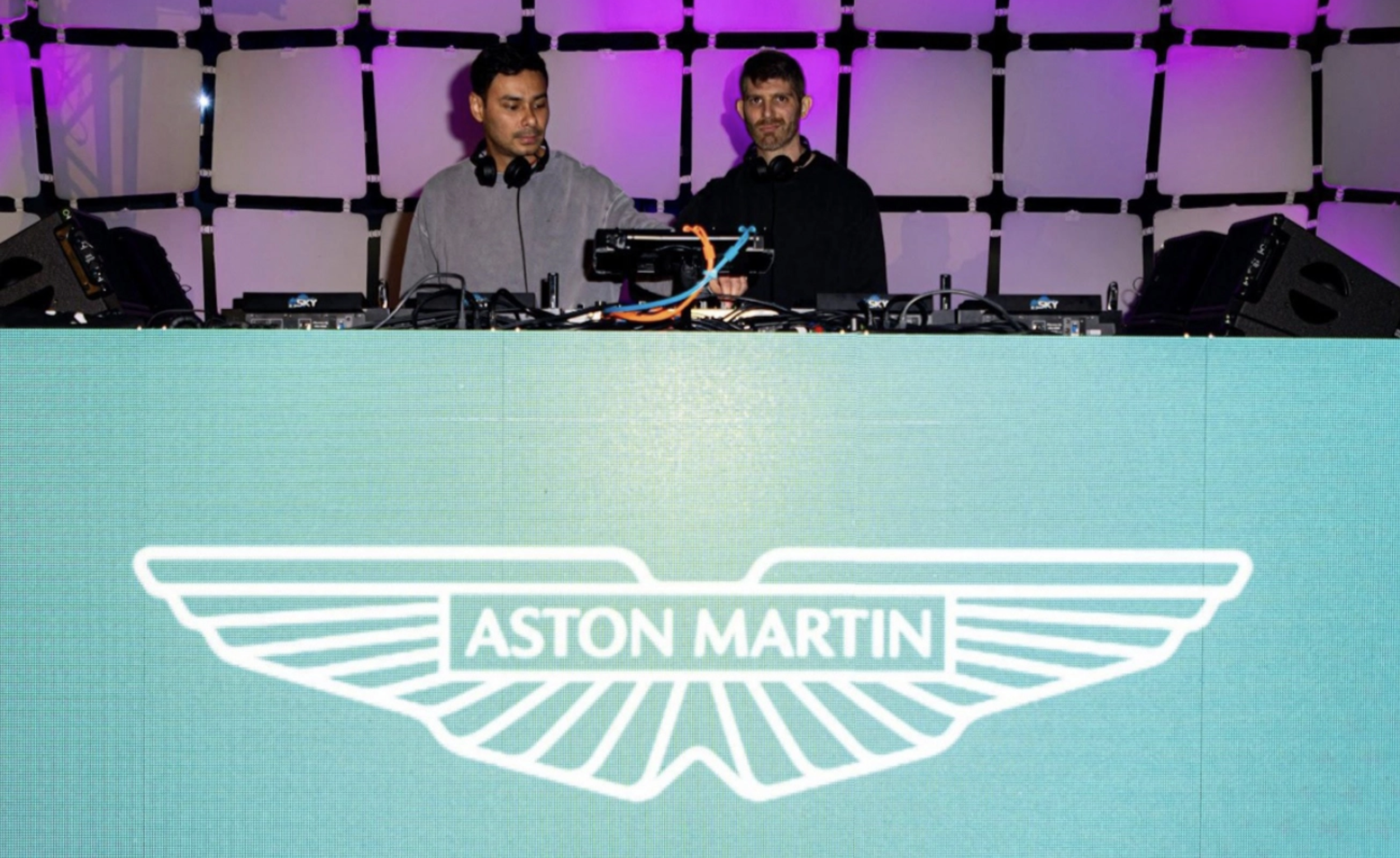 Two DJs or music producers performing at an event behind a large screen displaying the Aston Martin logo, with music equipment and speakers on stage, purple and pink lighting in the background.
