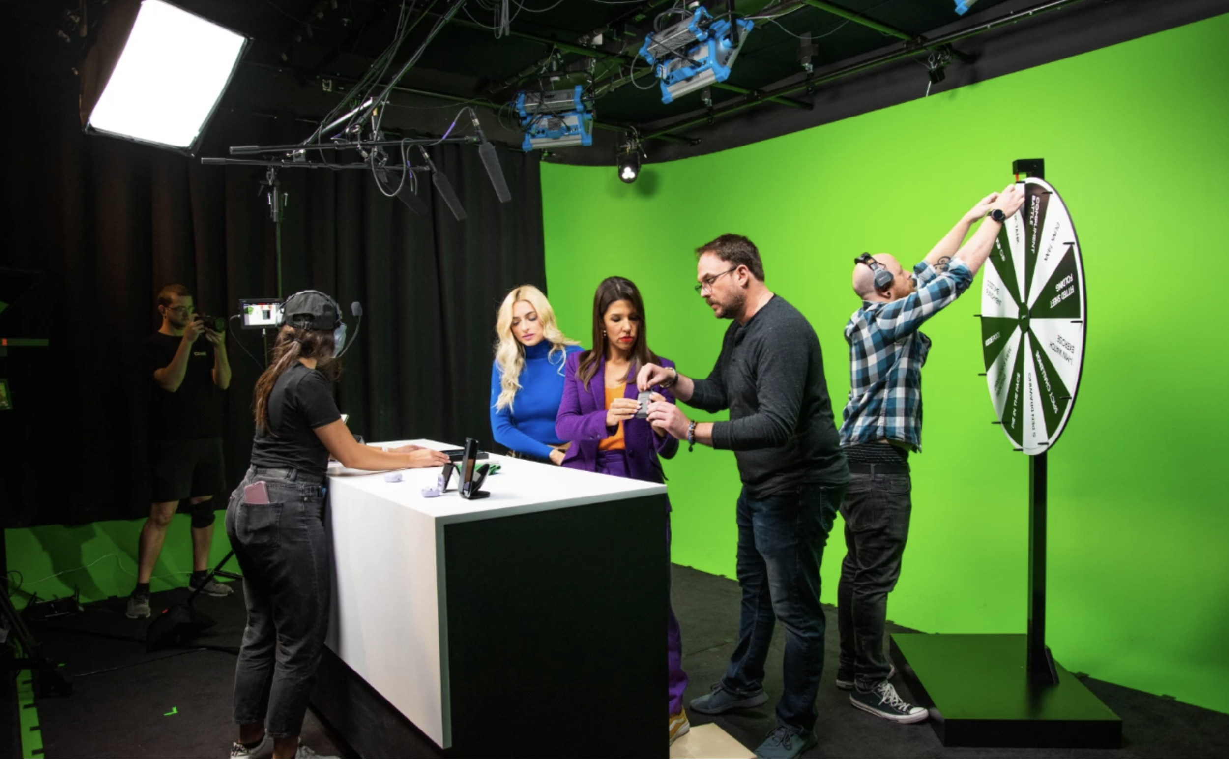 People filming with a green screen backdrop, a woman behind a white desk, and a spinning wheel with segments on a stand in a studio.