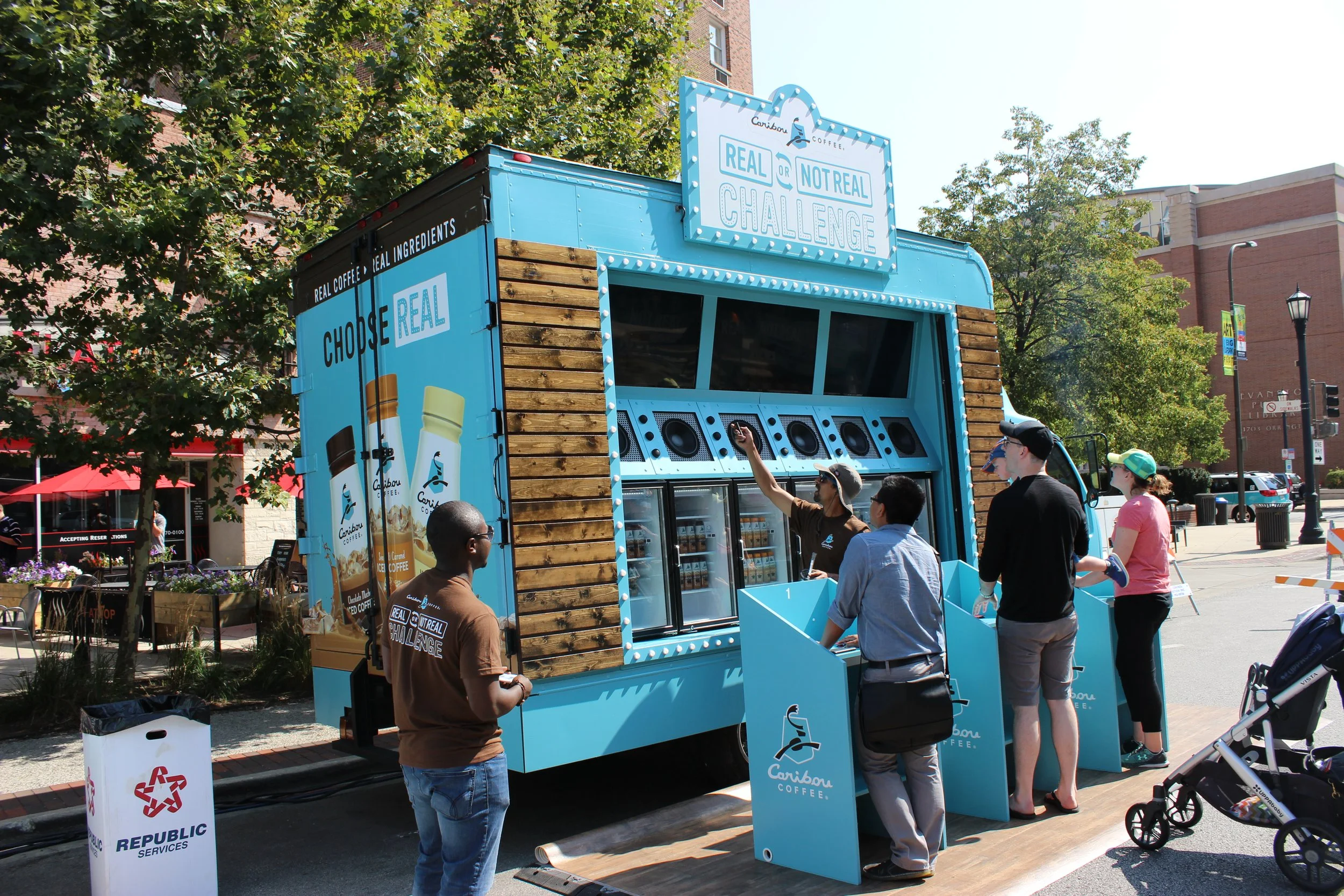 A blue coffee truck with a sign that reads 'Real or Not Real Challenge' and 'Choose Real' with images of bottled coffee drinks. Several people are in line at the truck, engaging with the staff. The scene is outdoors on a sunny day with trees and buildings in the background.