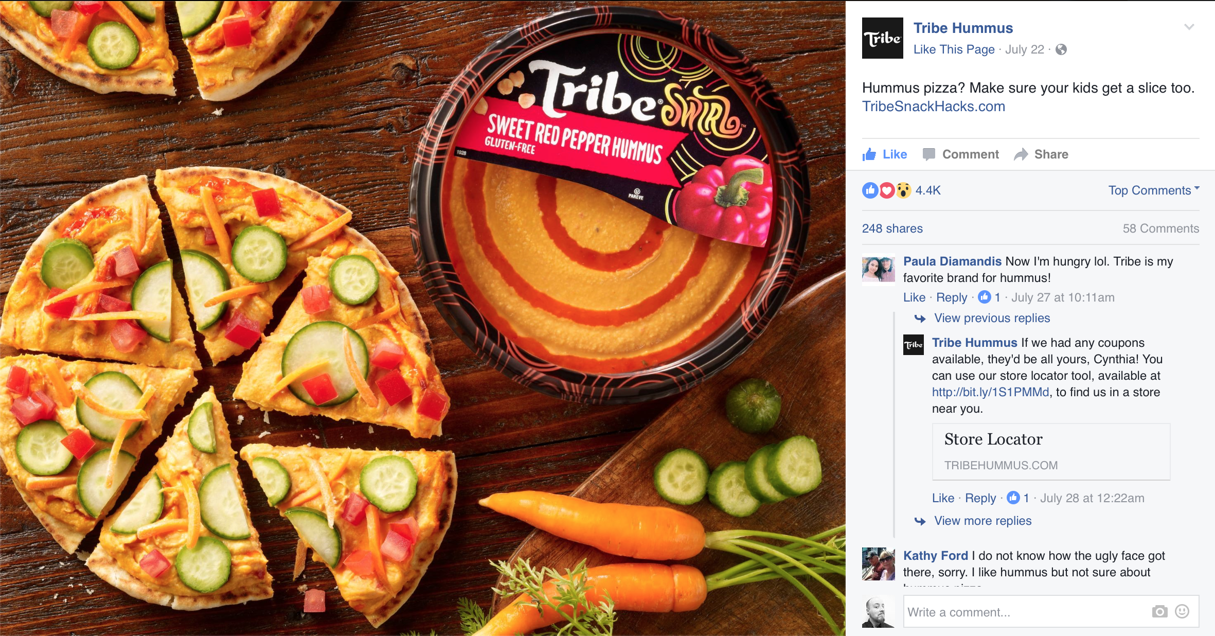 A package of Tribe Sweet Red Pepper Hummus next to a partially sliced veggie pizza topped with cucumbers, red bell peppers, and shredded cheddar cheese on a wooden surface, with whole carrots and sliced cucumbers nearby.