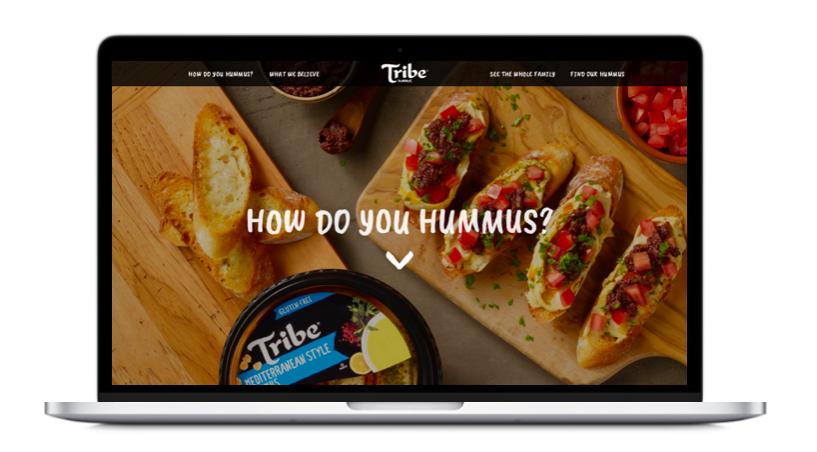 A laptop displaying a website with a wooden platter of baguette slices and topped toasts with colorful toppings, a container of hummus, and a bowl of chopped tomatoes, with text asking "How do you hummus?"