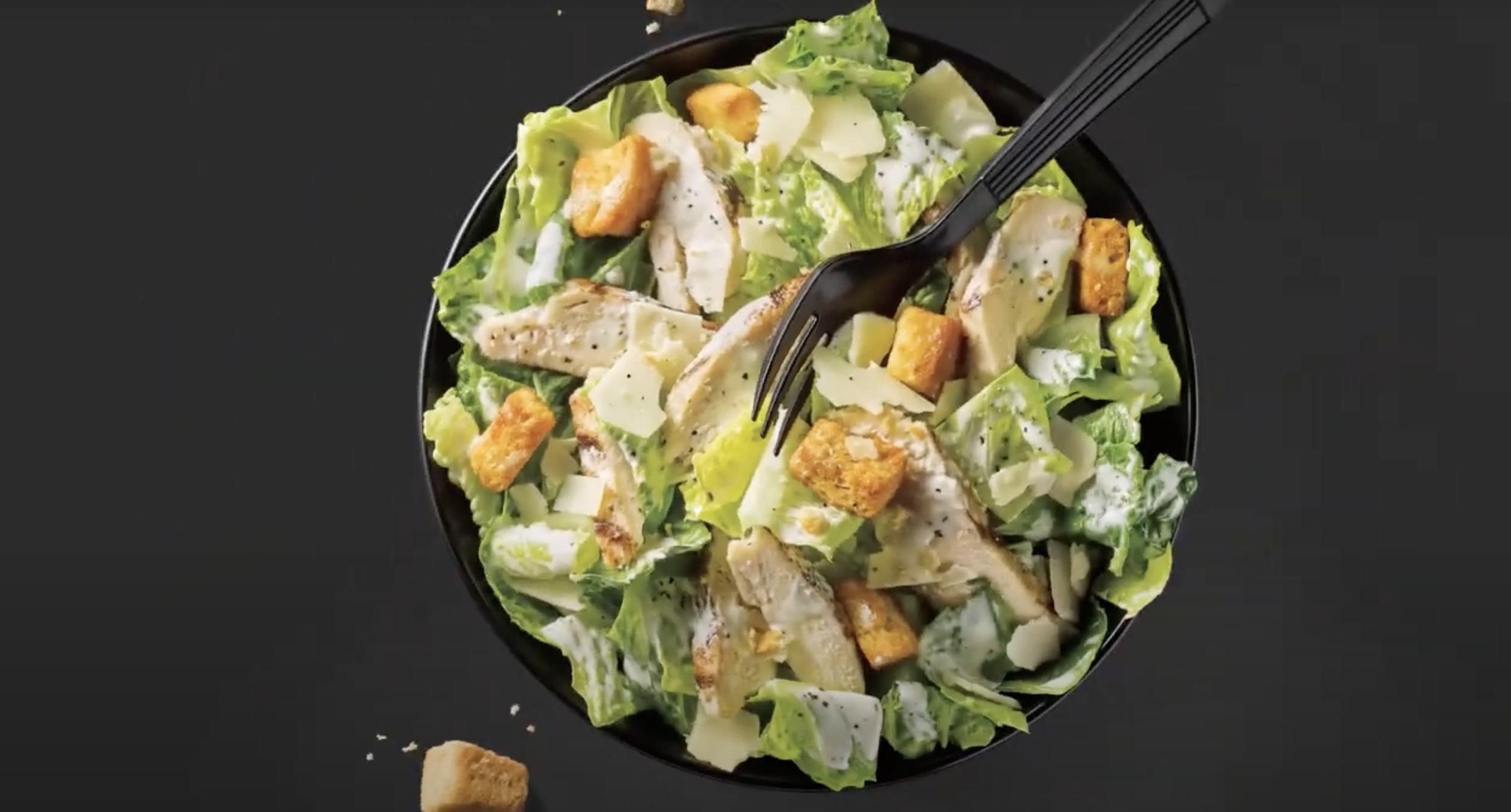 Caesar salad in a black bowl with lettuce, chicken, croutons, parmesan cheese, and black pepper, with a black fork on a black surface.