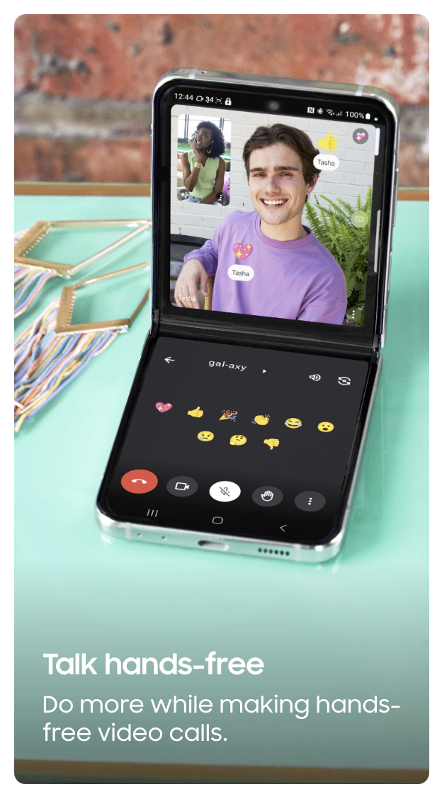 Smartphone open in a video call, showing a young man smiling and a young woman in a small inset, with a chat and emoji options visible on the screen, placed on a teal table with fabric and gold rings nearby.