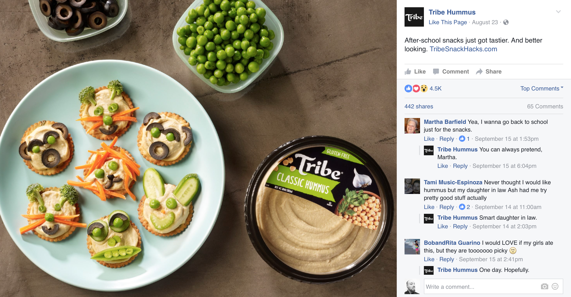 Individual crackers decorated with hummus, vegetables, and olives resembling animal faces, served on a light blue plate, with bowls of green peas and black olives nearby, and a container of Tribe classic hummus.