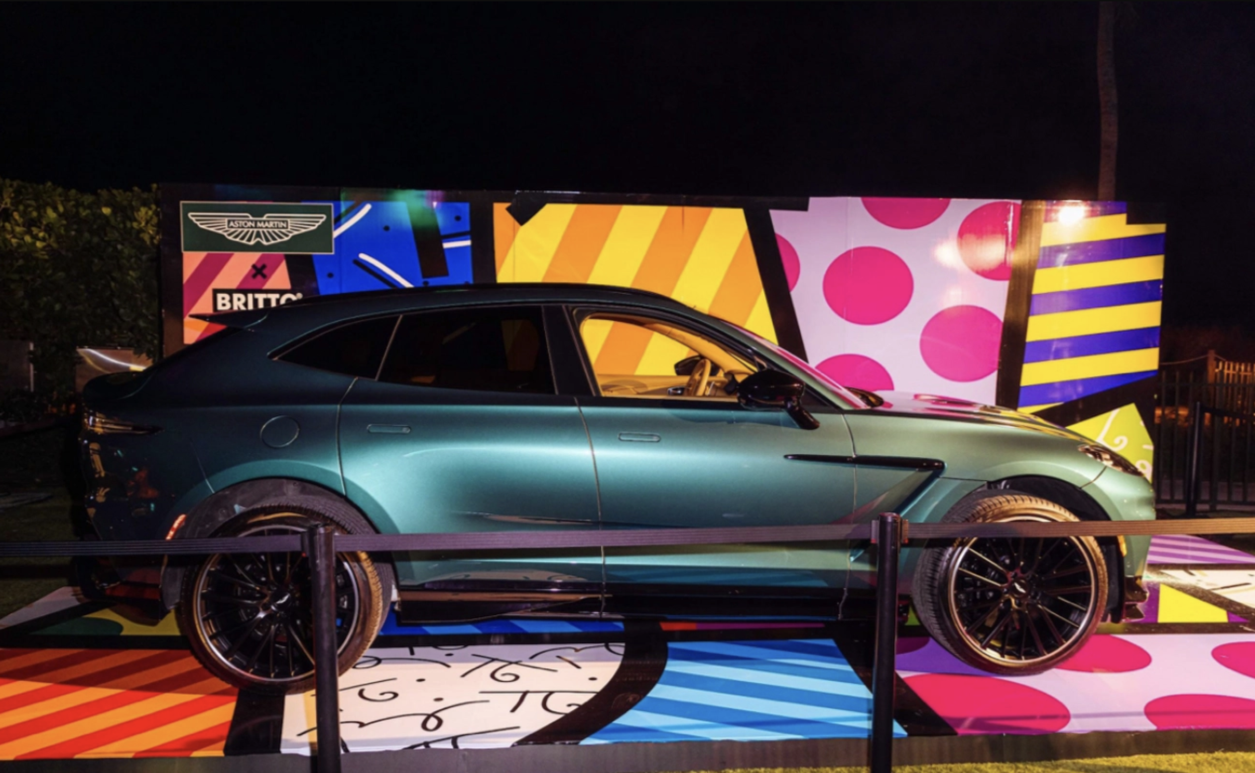 A luxury teal-colored car displayed at an event with colorful geometric background and lighting.
