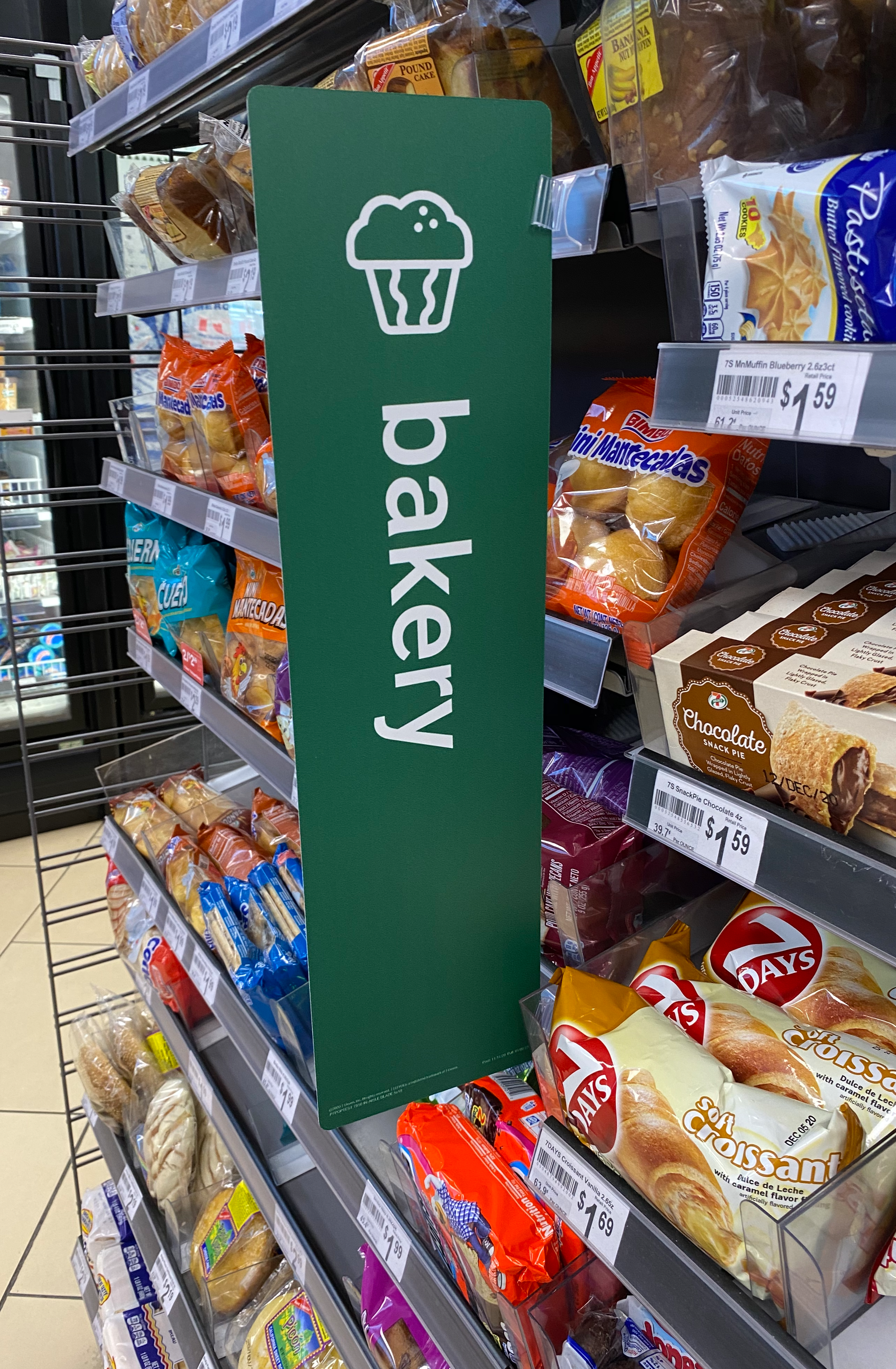 Grocery store shelves filled with various baked goods, including packaged bread and sweet snacks. A green shopping cart sign with a cupcake icon and the word 'bakercy' is hanging on the shelf.