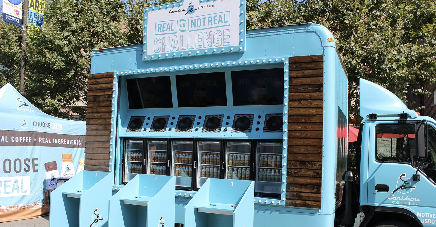 A blue mobile coffee truck with a wooden panel accent and a large sign on top reading 'Real or Not Real Challenge' by Caribou Coffee, featuring windows with refrigerated compartments and three blue kiosks in front.