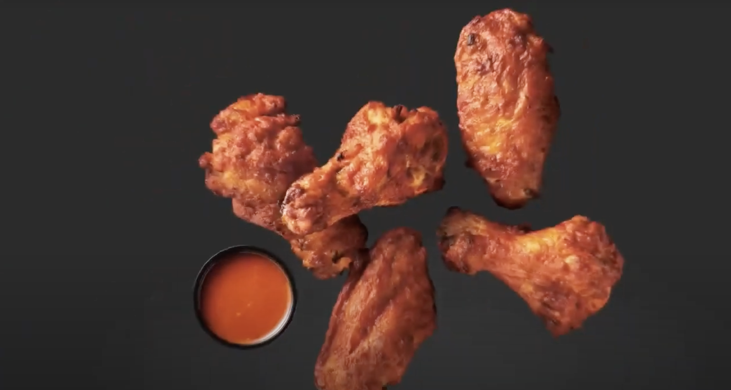 Four pieces of breaded, fried chicken wings with a small cup of spicy sauce on the side, on a black background.