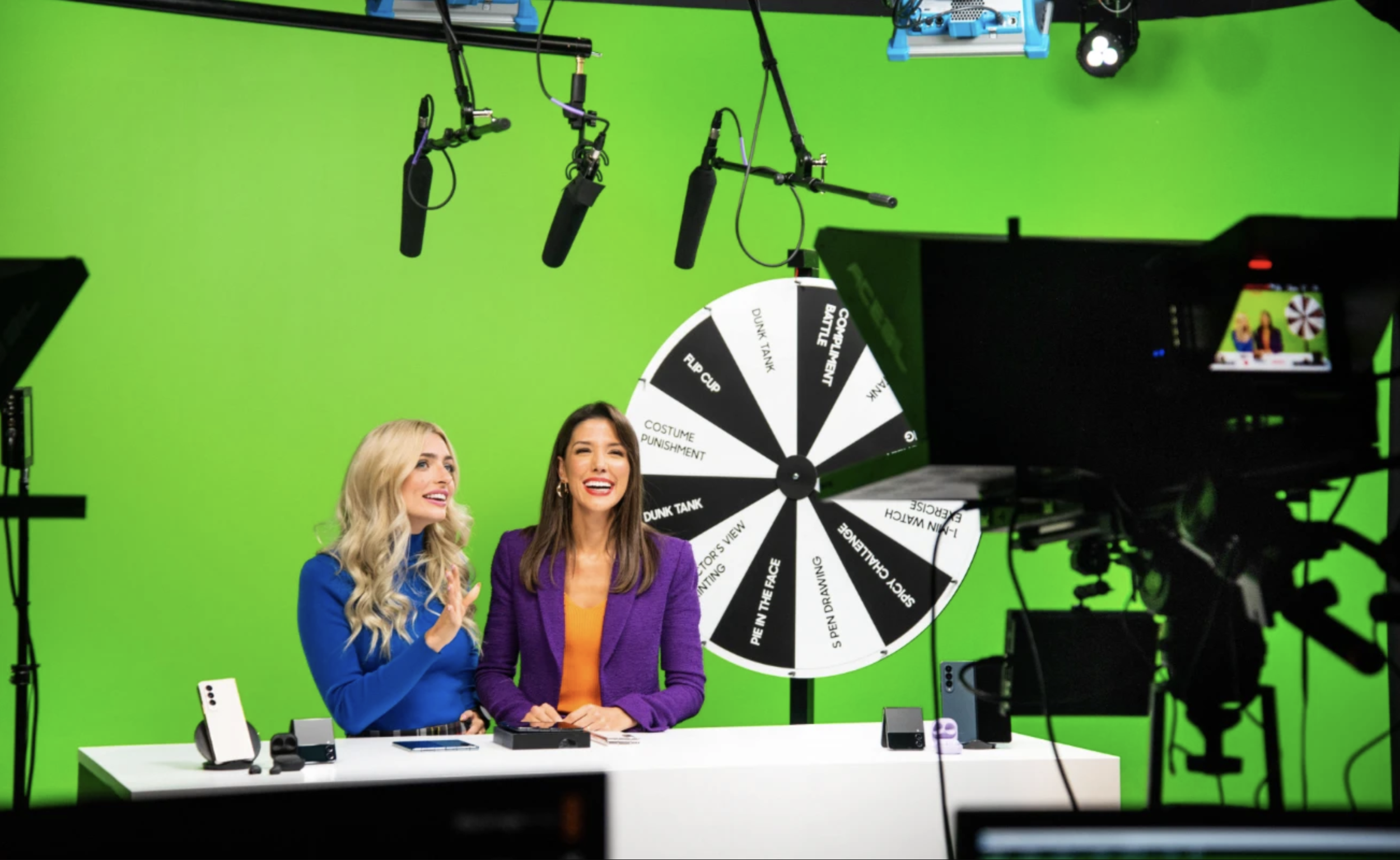 Two women sitting at a white table, smiling and talking, in a brightly lit TV studio with a large green screen background. There is a spinning wheel with various sections behind them, surrounded by professional filming equipment including cameras, microphones, and lighting.